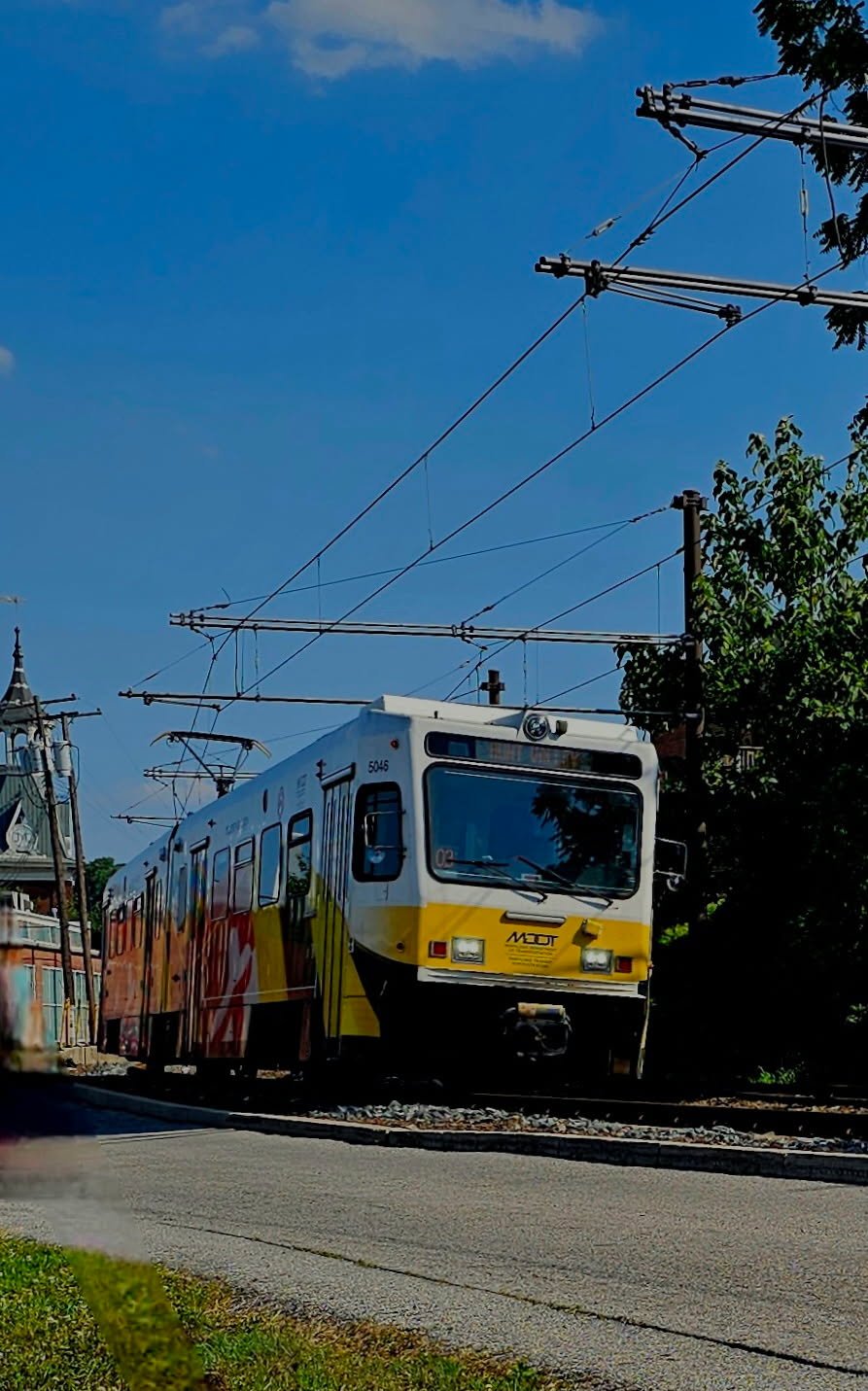A streetcar or tram on a city street with overhead electric wires, trees, and buildings in the background on a sunny day.