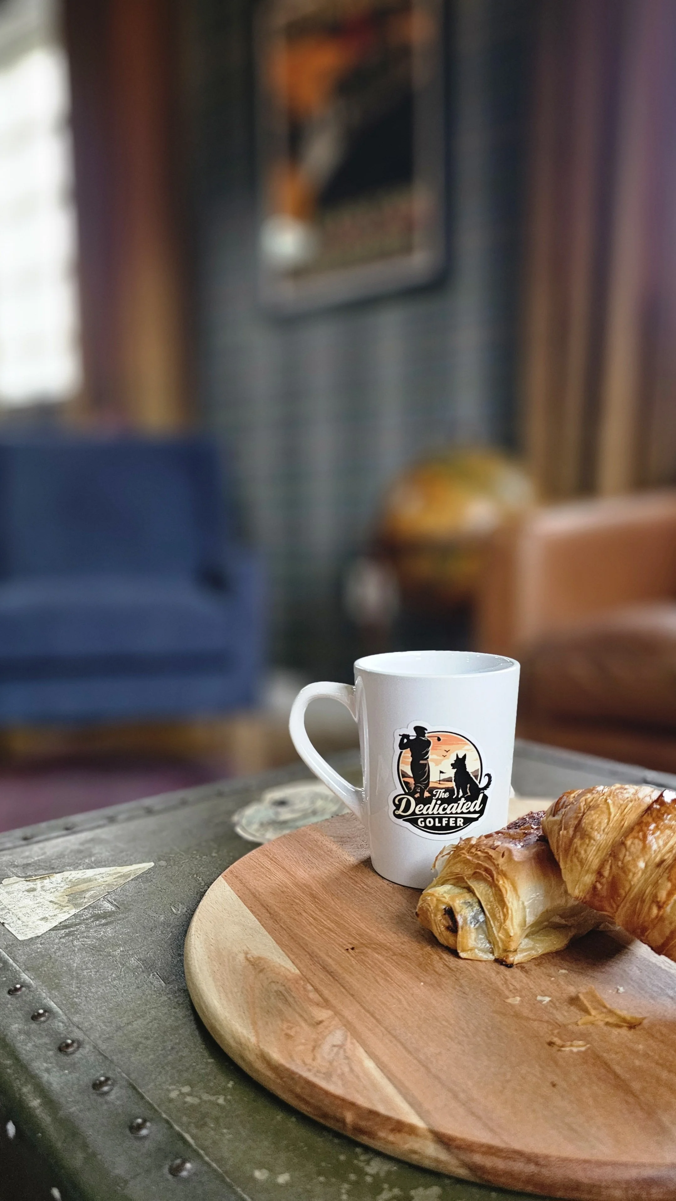 Coffee mug with a golf logo and a croissant on a wooden serving board, placed on a table.