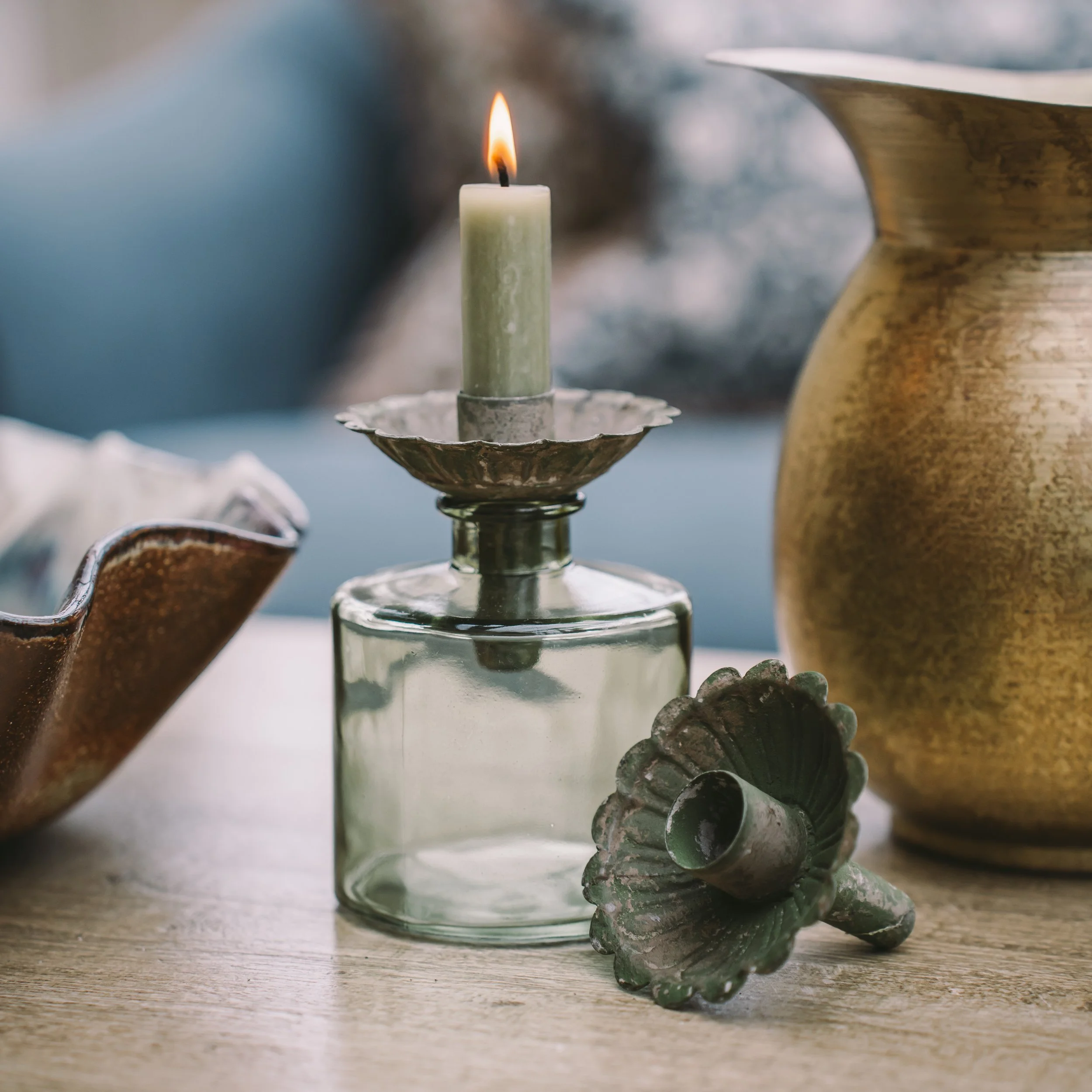 A lit white candle in a glass holder on a wooden surface, with decorative ceramic objects nearby and a blurred background.