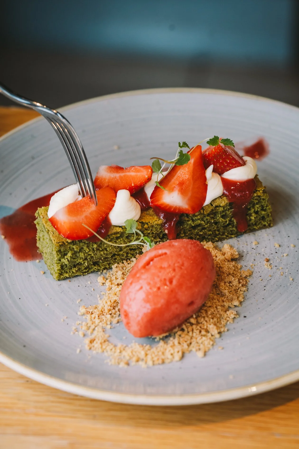 A slice of green matcha-flavored cake topped with sliced strawberries, whipped cream, microgreens, and strawberry sauce on a gray plate, with a scoop of pink sorbet and crushed nuts on the side.