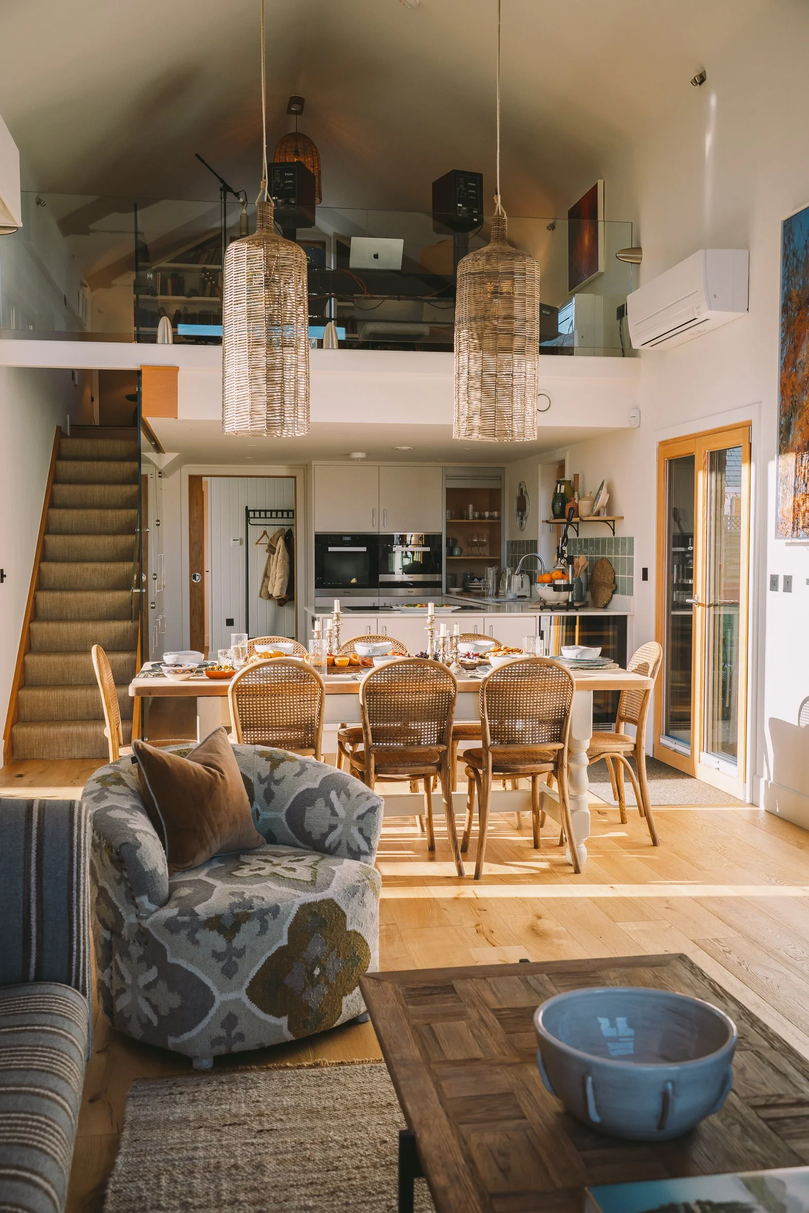 A dining area with a long wooden table set with dinnerware, surrounded by wooden chairs. A patterned armchair and a striped sofa with pillows are in the foreground. In the background, a kitchen with white cabinets, a black oven, and open shelving is 