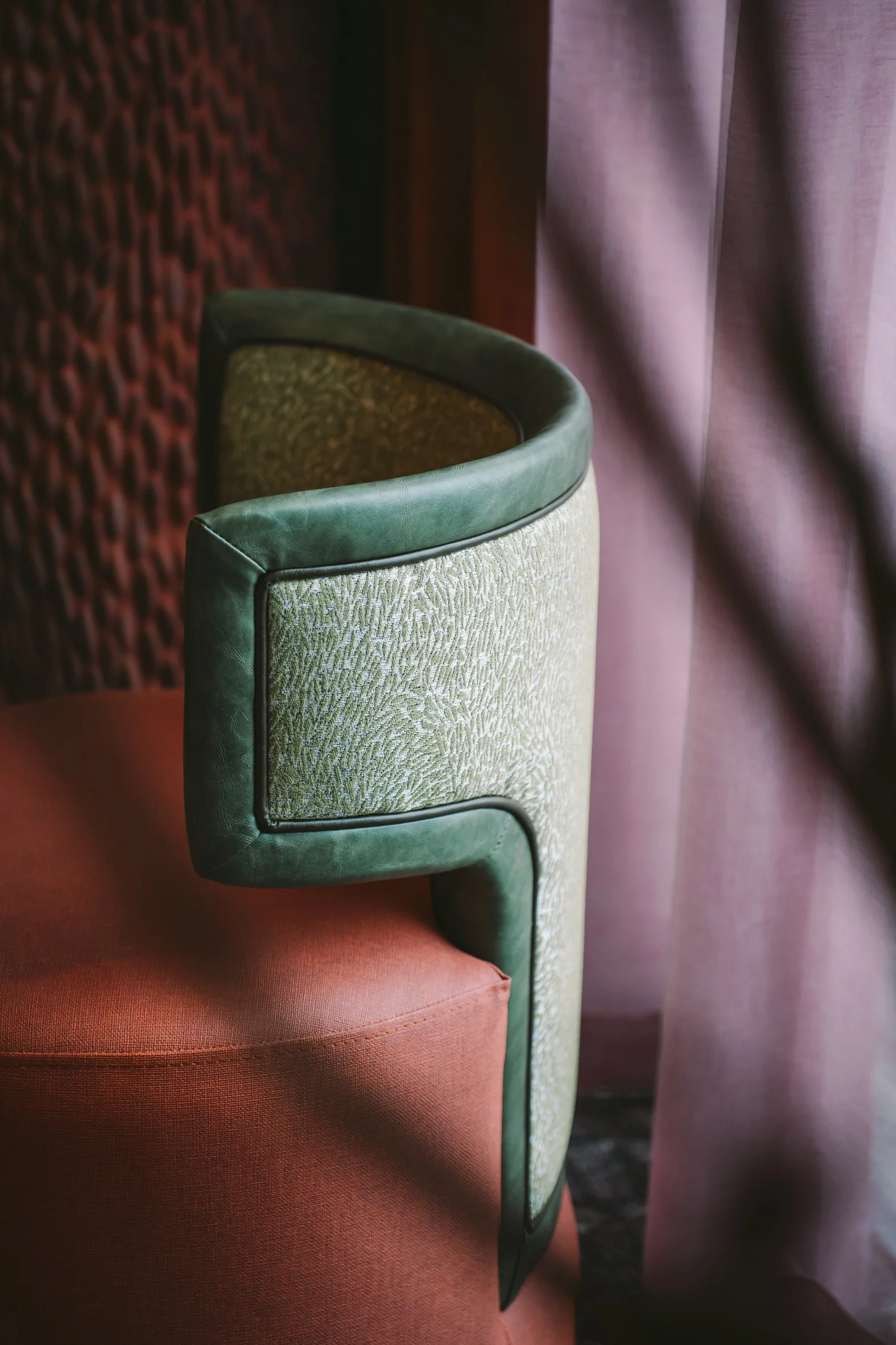 Close-up of a vintage-style armchair with green velvet upholstery and patterned fabric, situated next to a purple curtain in a dimly lit room.