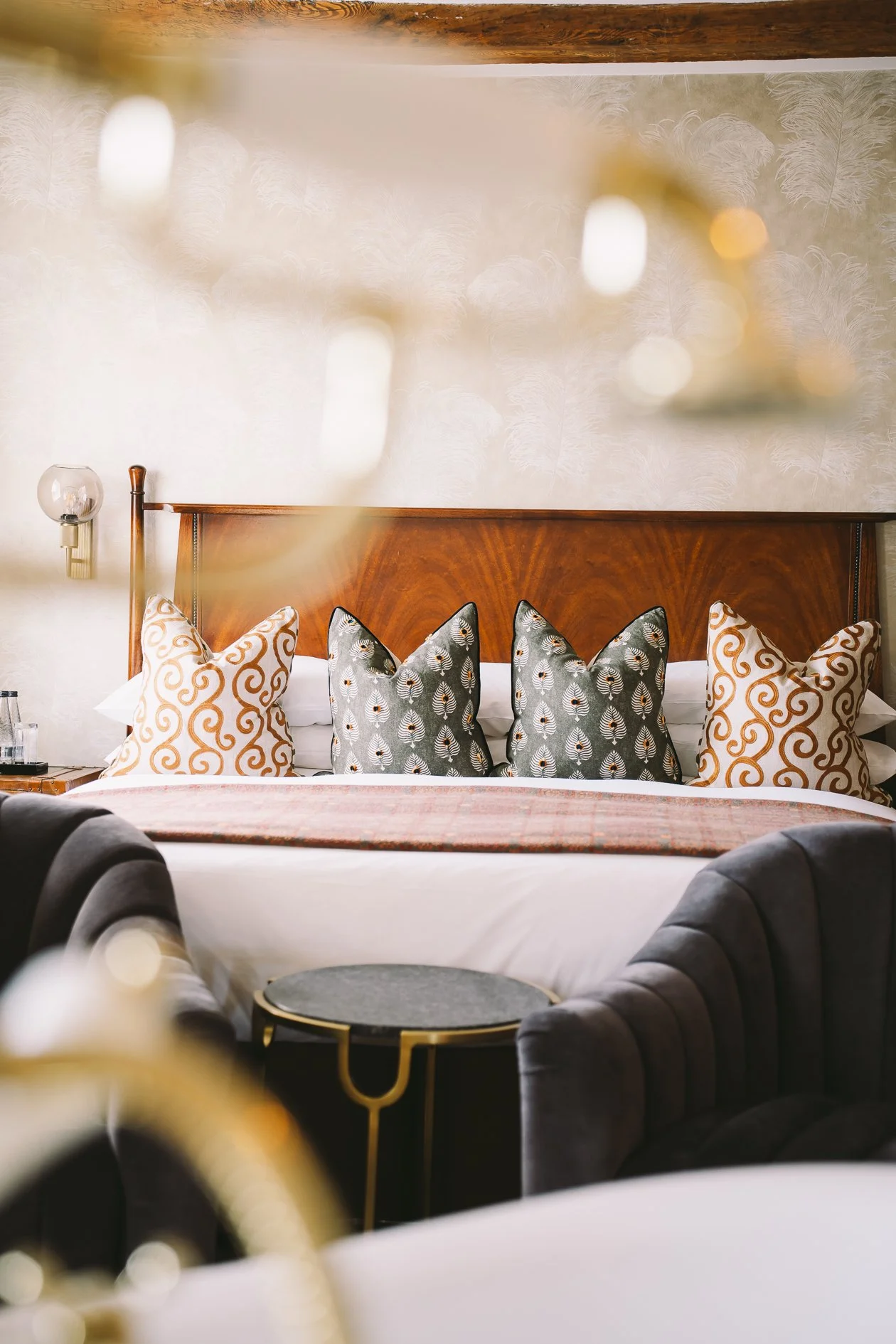 A bed with decorative pillows, wooden headboard, a wall-mounted lamp, and blurred foreground elements in a stylish bedroom.