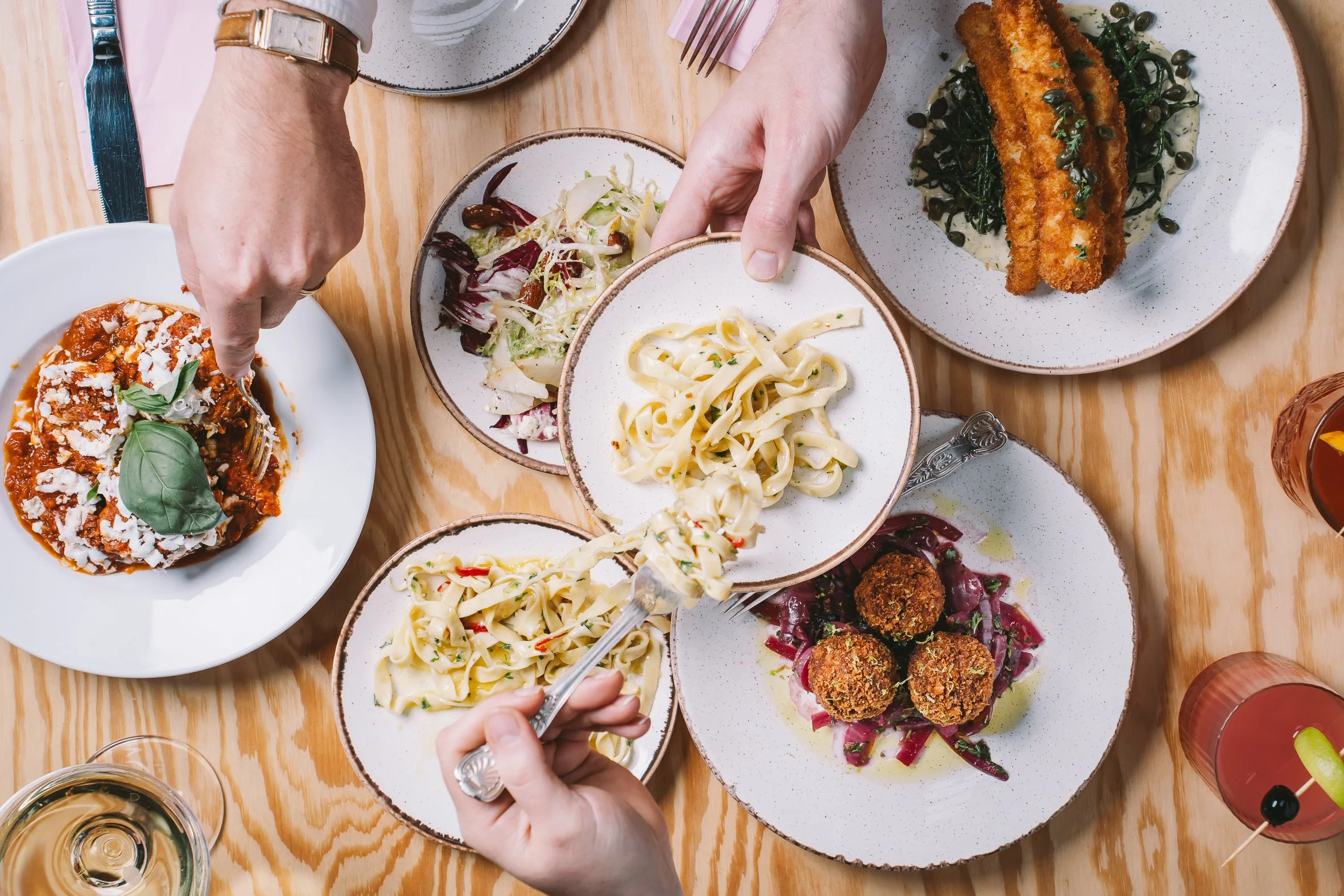 A top-down view of a meal with various dishes including pasta, salad, fried fish with greens, stuffed meatballs, and a dessert, on a wooden table with wine glasses and utensils.