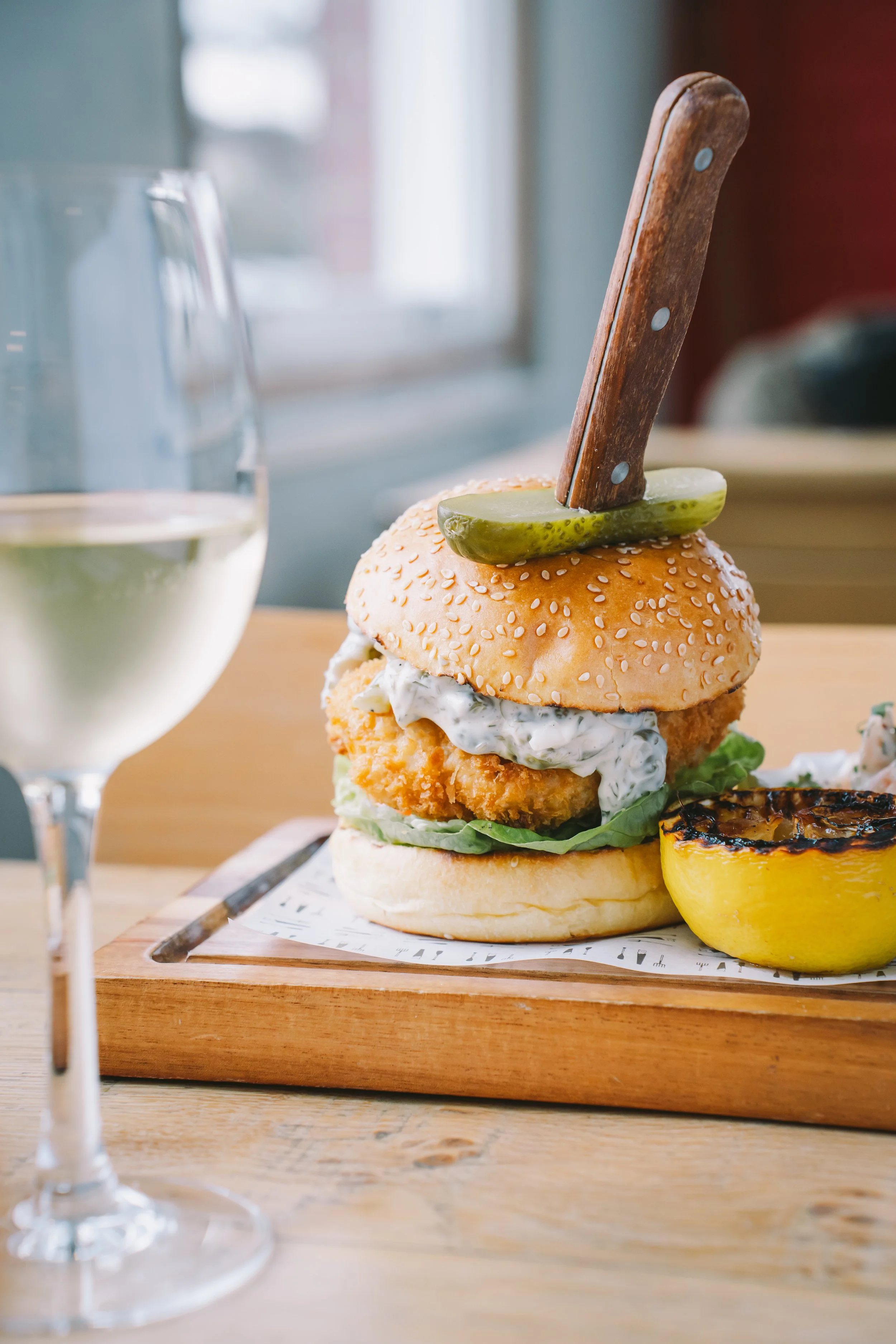 A close-up of a crispy fried chicken sandwich with lettuce, mayonnaise, and a sesame seed bun, topped with a pickle spear and a knife stuck upright through the pickle. A glass of white wine and a grilled half lemon are visible on the table.