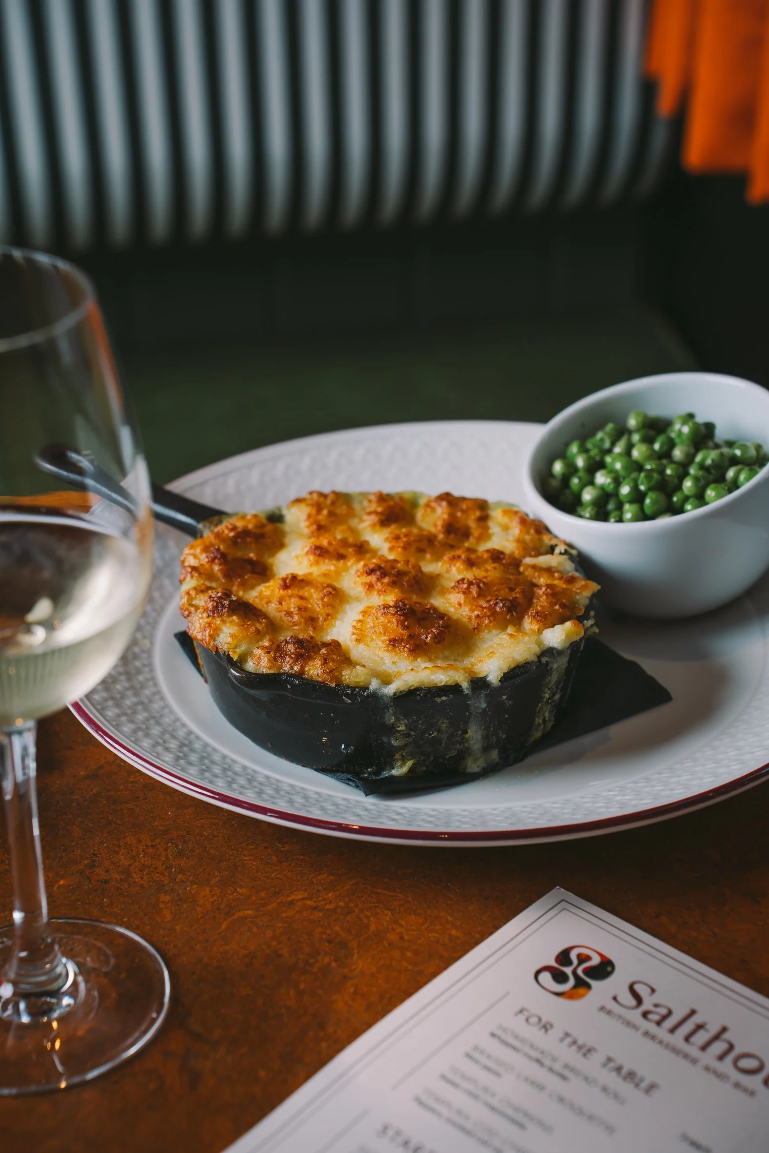 A plate of baked macaroni and cheese with a golden-brown top, a bowl of green peas, a glass of white wine, a wine glass, and a menu on a wooden table.