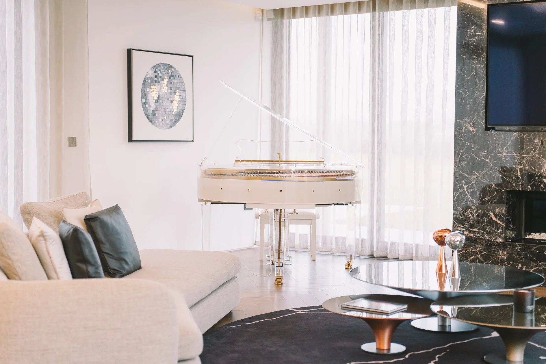 Modern living room with a white grand piano near large windows with sheer curtains, a beige sofa with black and white pillows, a black marble fireplace with a mounted flat-screen TV, and a round black coffee table with decorative vases.