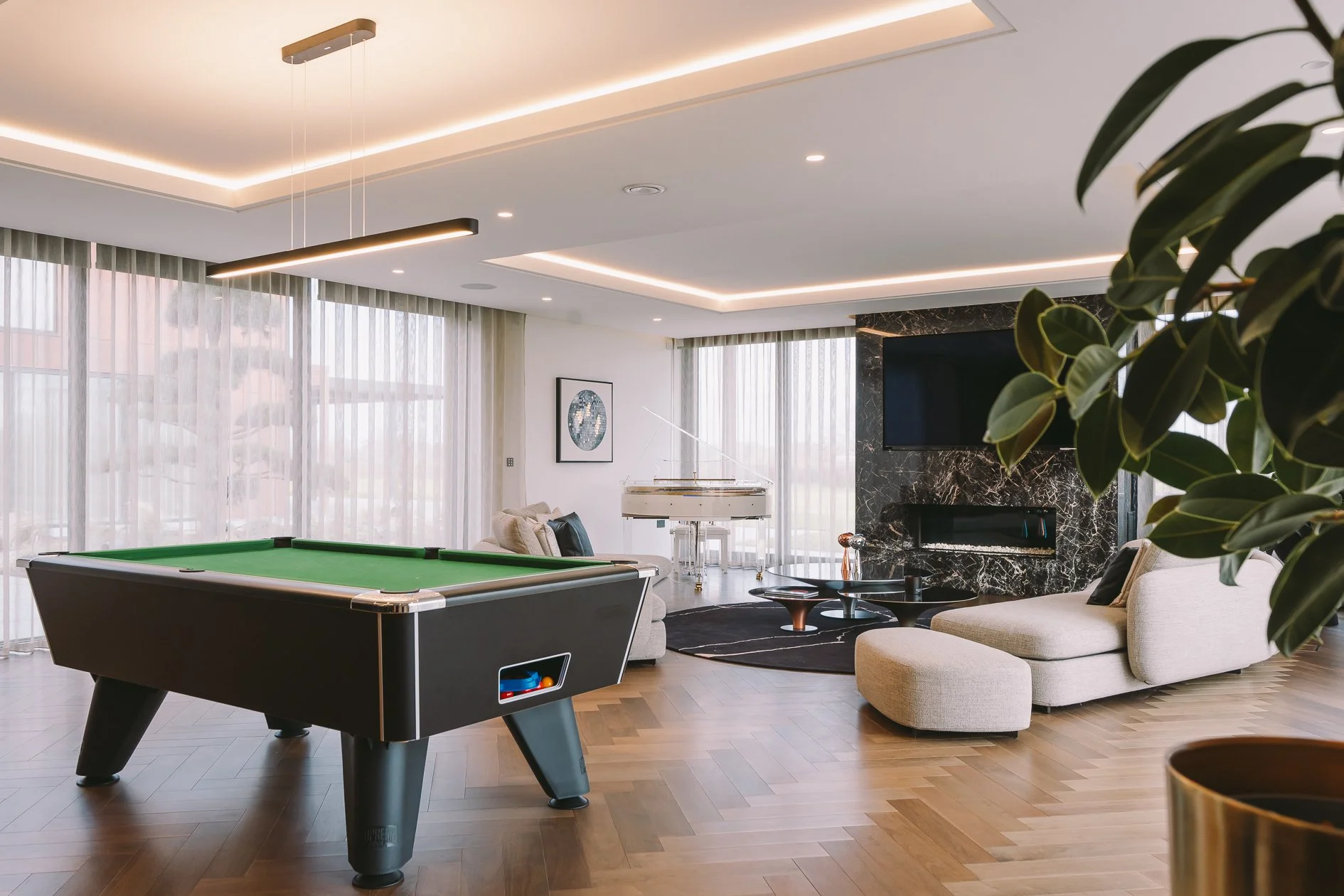 Modern living room with a pool table, white sofa, black fireplace with marble surround, flat screen TV, and a grand white piano near large windows with sheer curtains, decorative artwork, and indoor plants.