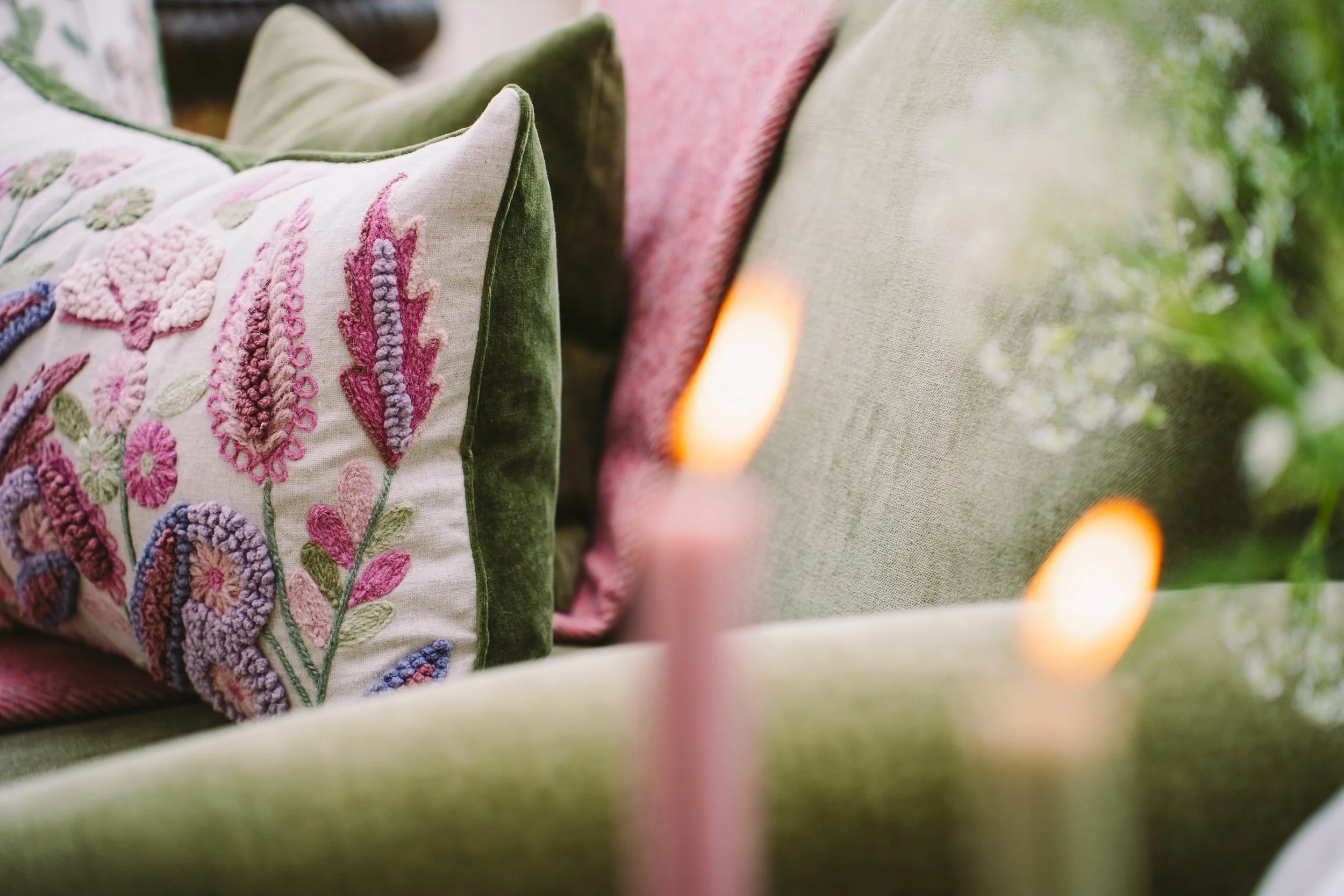Close-up of decorative pillows on outdoor furniture, featuring intricate embroidery and pastel colors, with greenery in the background.