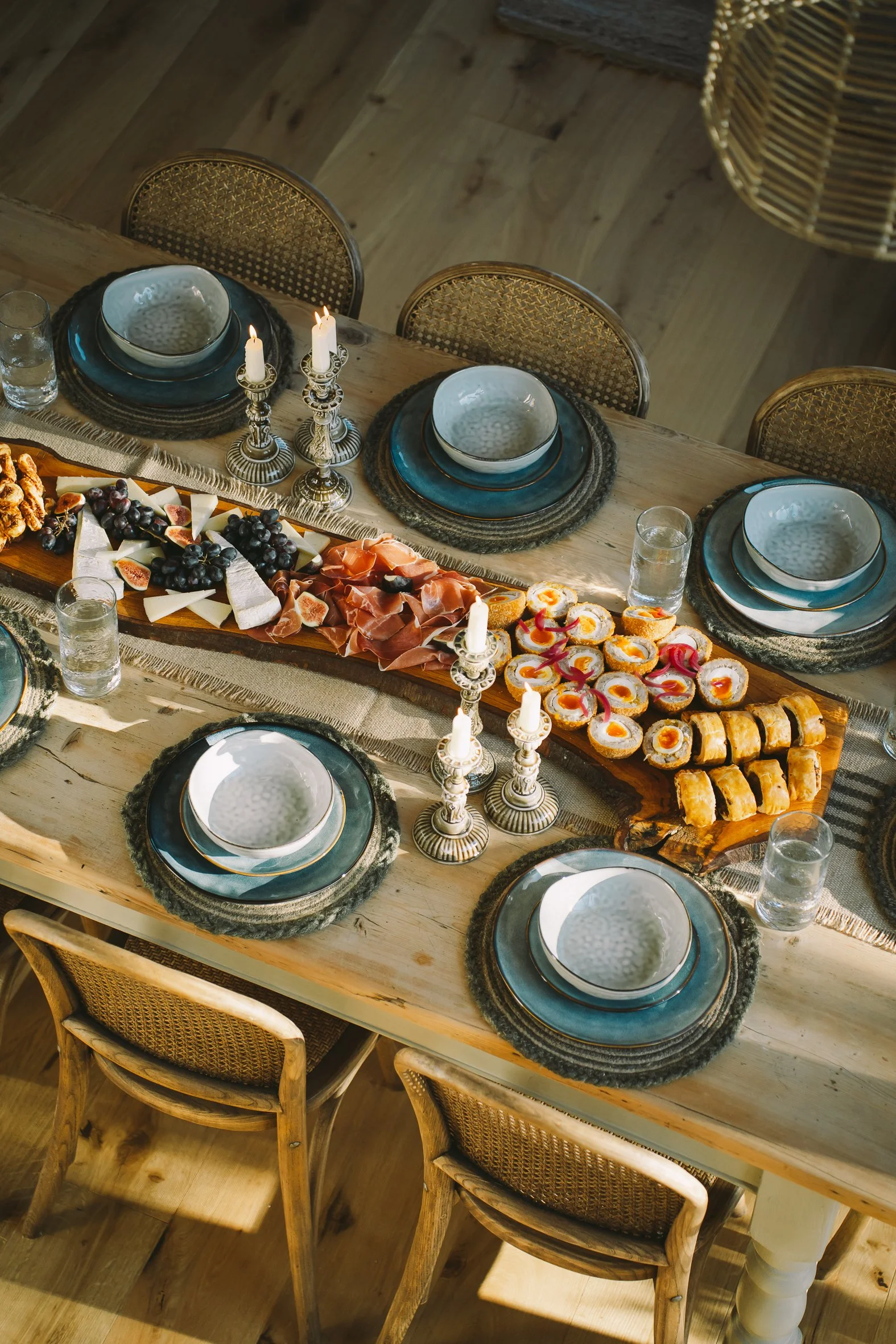 A dining table decorated for a meal with plates, glasses, and a large platter of cheese, grapes, meats, and sushi, illuminated by candles.