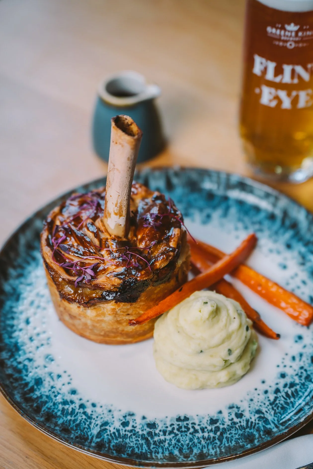 A plate with a stuffed Yorkshire pudding topped with gravy and microgreens, served with mashed potatoes, roasted carrots, and a dollop of herb butter. In the background, a bottle of pale ale and a small blue pitcher are visible.