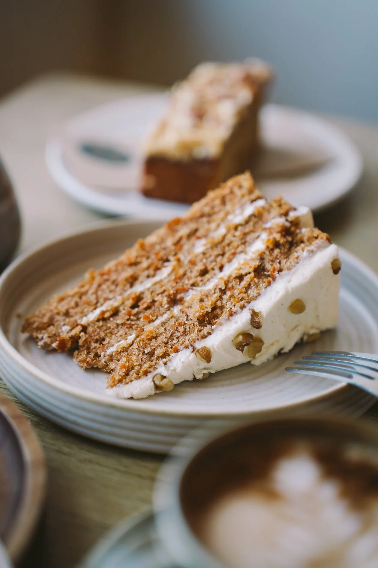 A slice of carrot cake with cream cheese frosting on a beige plate, with a cup of coffee and a blurred slice of cake on a white plate in the background.