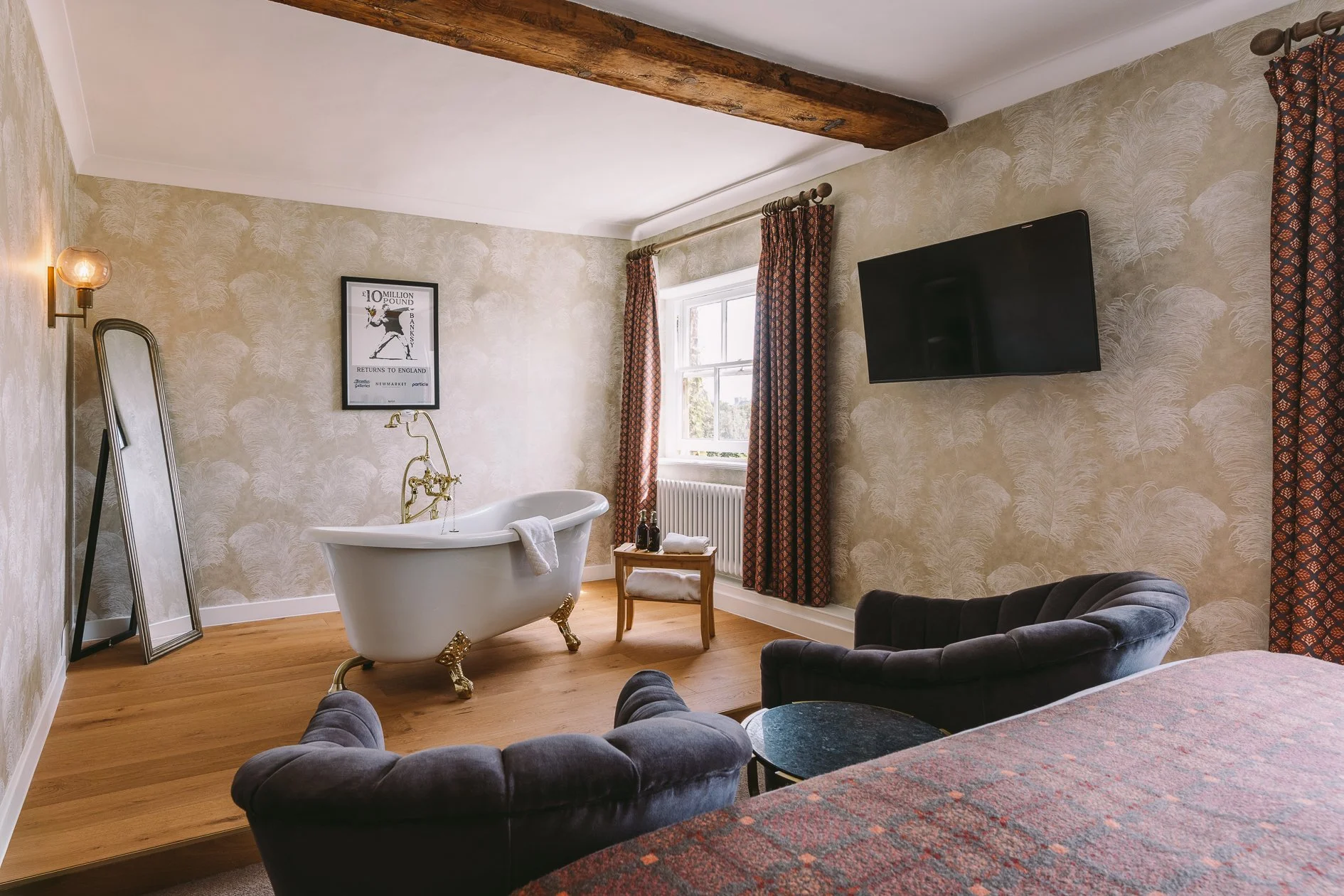 A cozy living room with a vintage bathtub, a black velvet armchair, a wall-mounted TV, framed artwork, and curtains on a window, with wooden floors and a rustic ceiling beam.