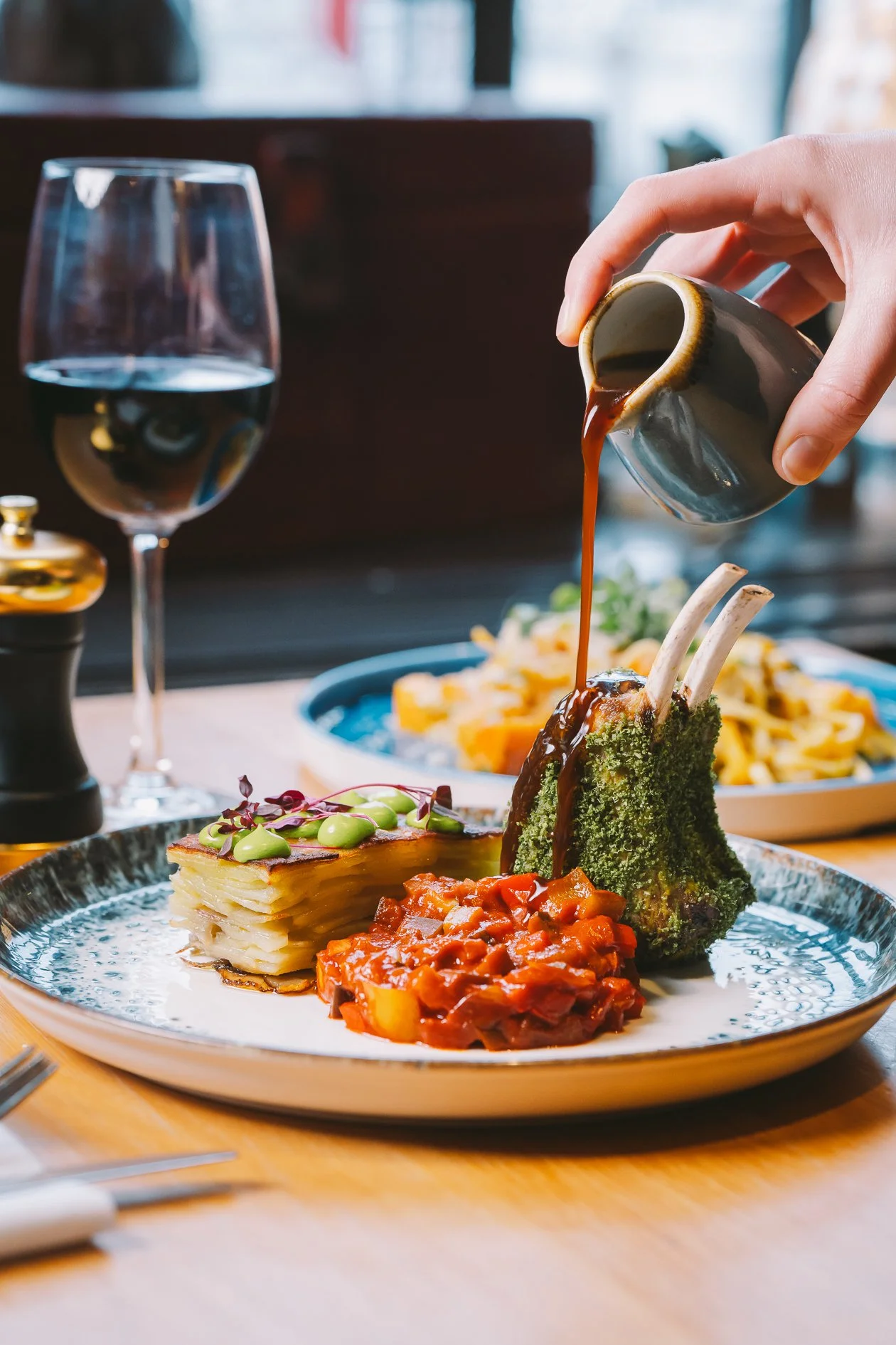 A hand pouring brown gravy over a plate of food including layered potato casserole, tomato-based stew, and a baked fish with grilled vegetables, with a glass of water and a glass salt shaker on the table.