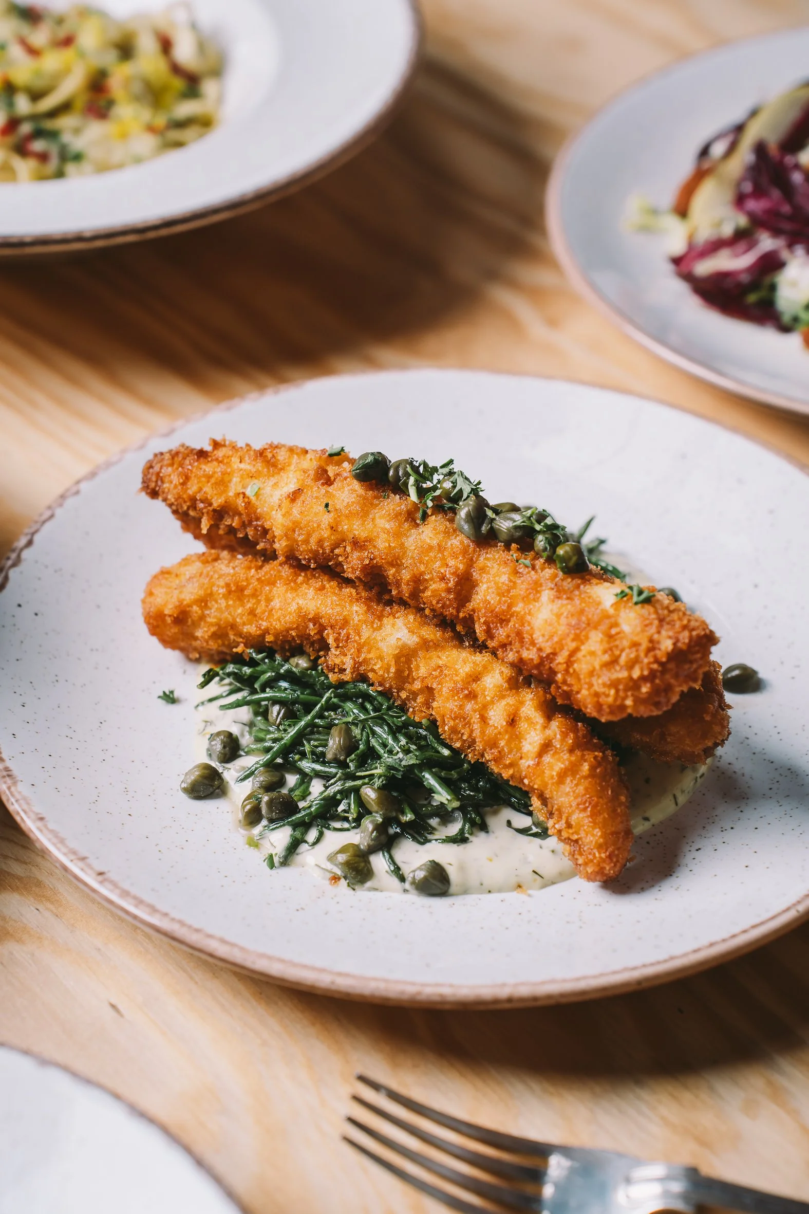 Fried fish fillets garnished with herbs and served on a plate with greens and capers, with additional dishes and cutlery on a wooden table.