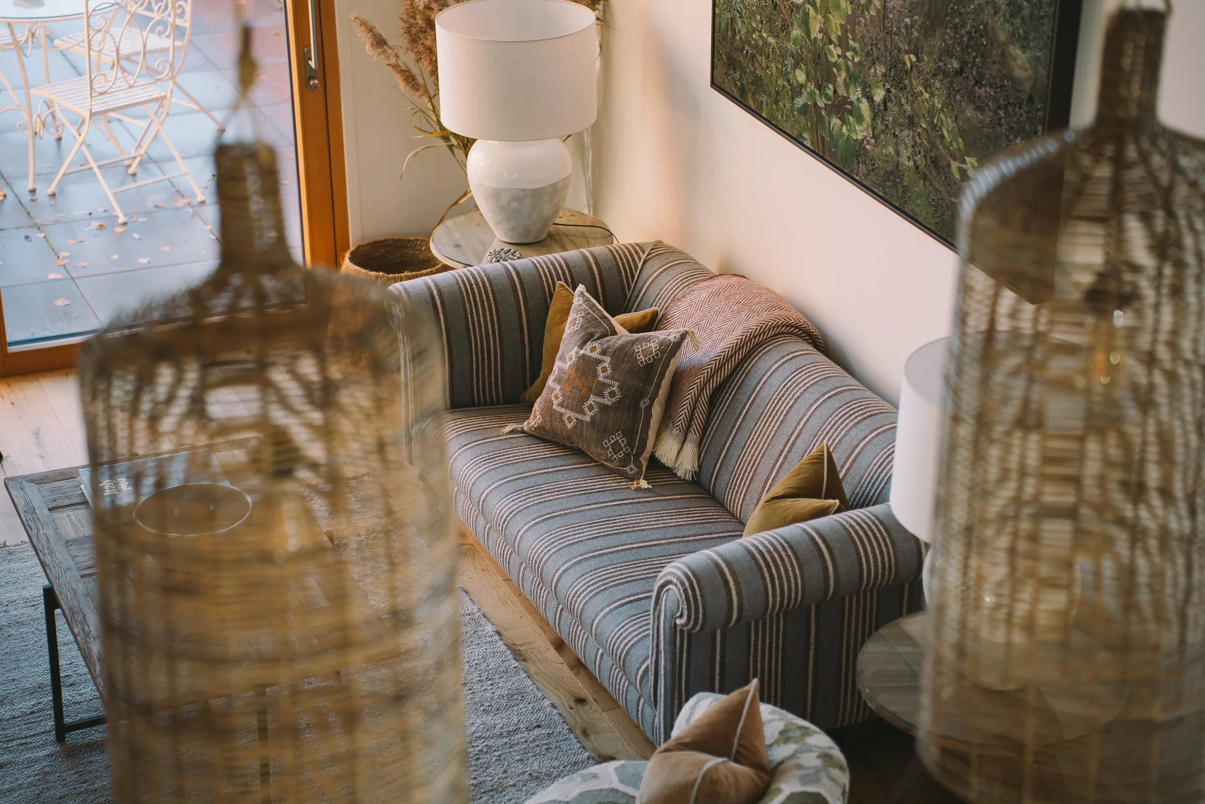 Living room with a striped sofa, decorative pillows, a side table with a lamp, and a large wall-mounted monitor showing outdoor greenery. The room has a wooden floor and a sliding glass door leading to a patio with outdoor chairs.