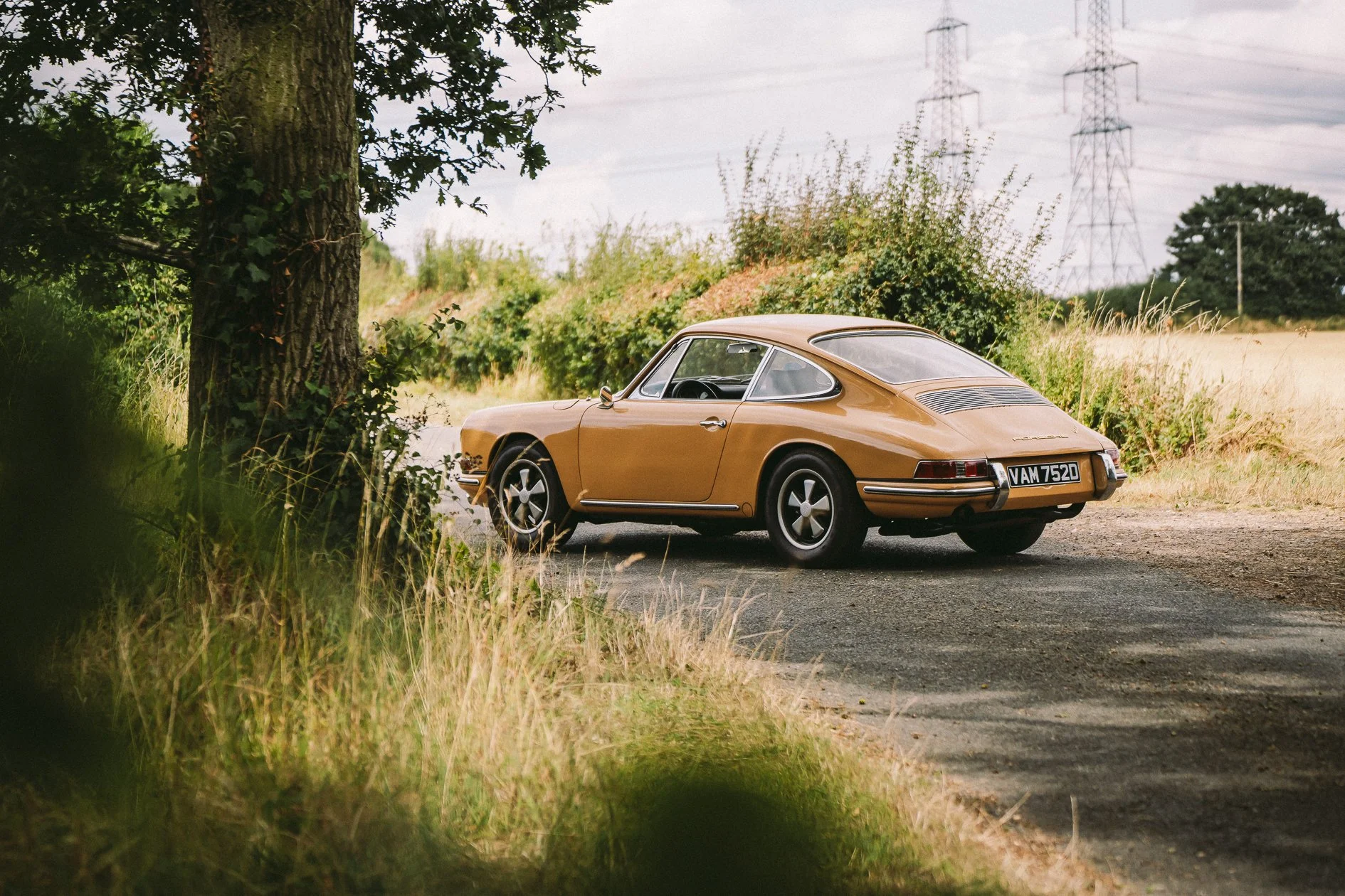A vintage brown Porsche car parked on a rural roadside with grass, trees, and power lines in the background.