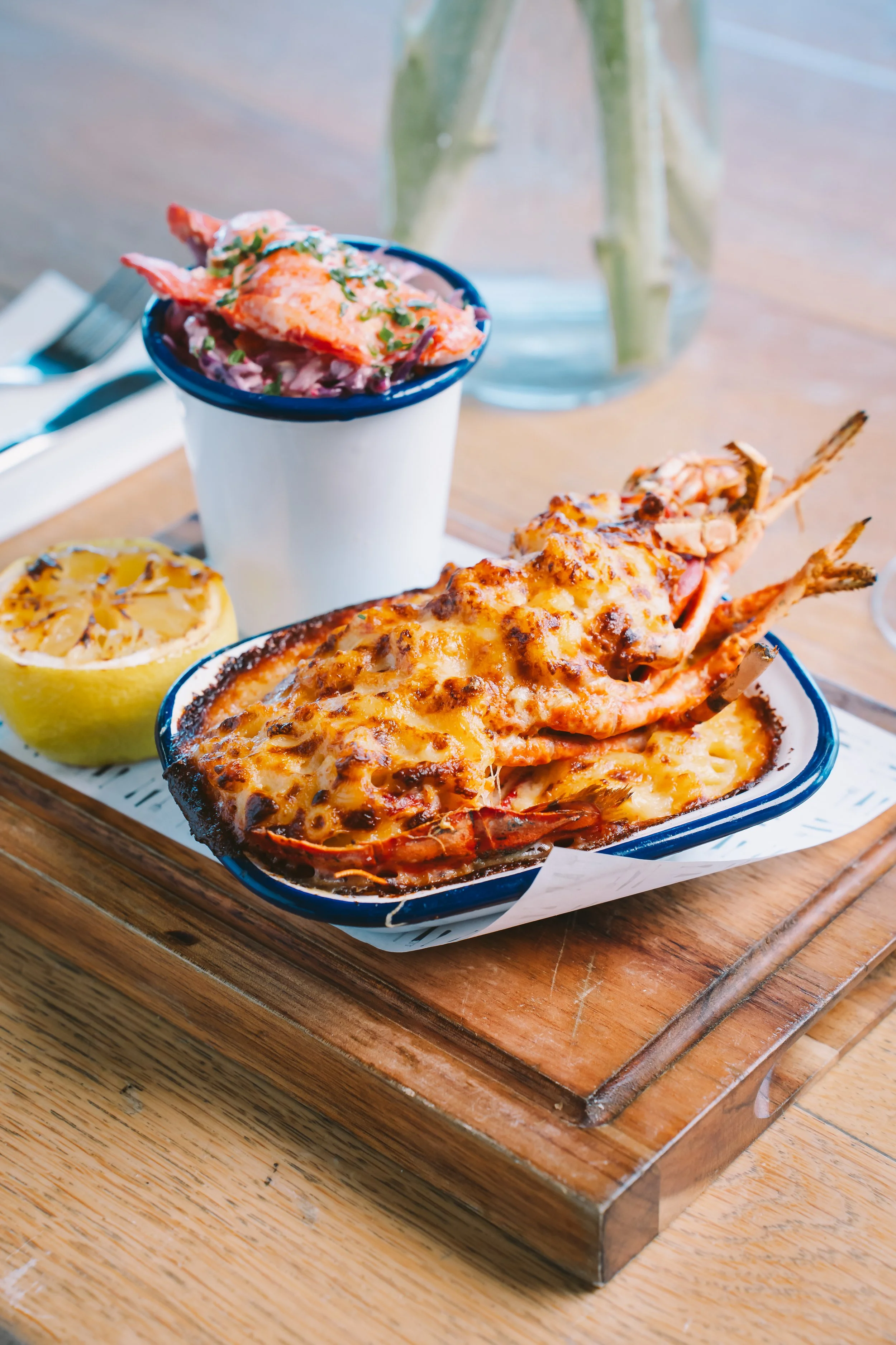 A seafood dish with baked lobster topped with cheese, served on a blue-rimmed dish, accompanied by a lemon half and a small bowl of salad, on a wooden serving tray.