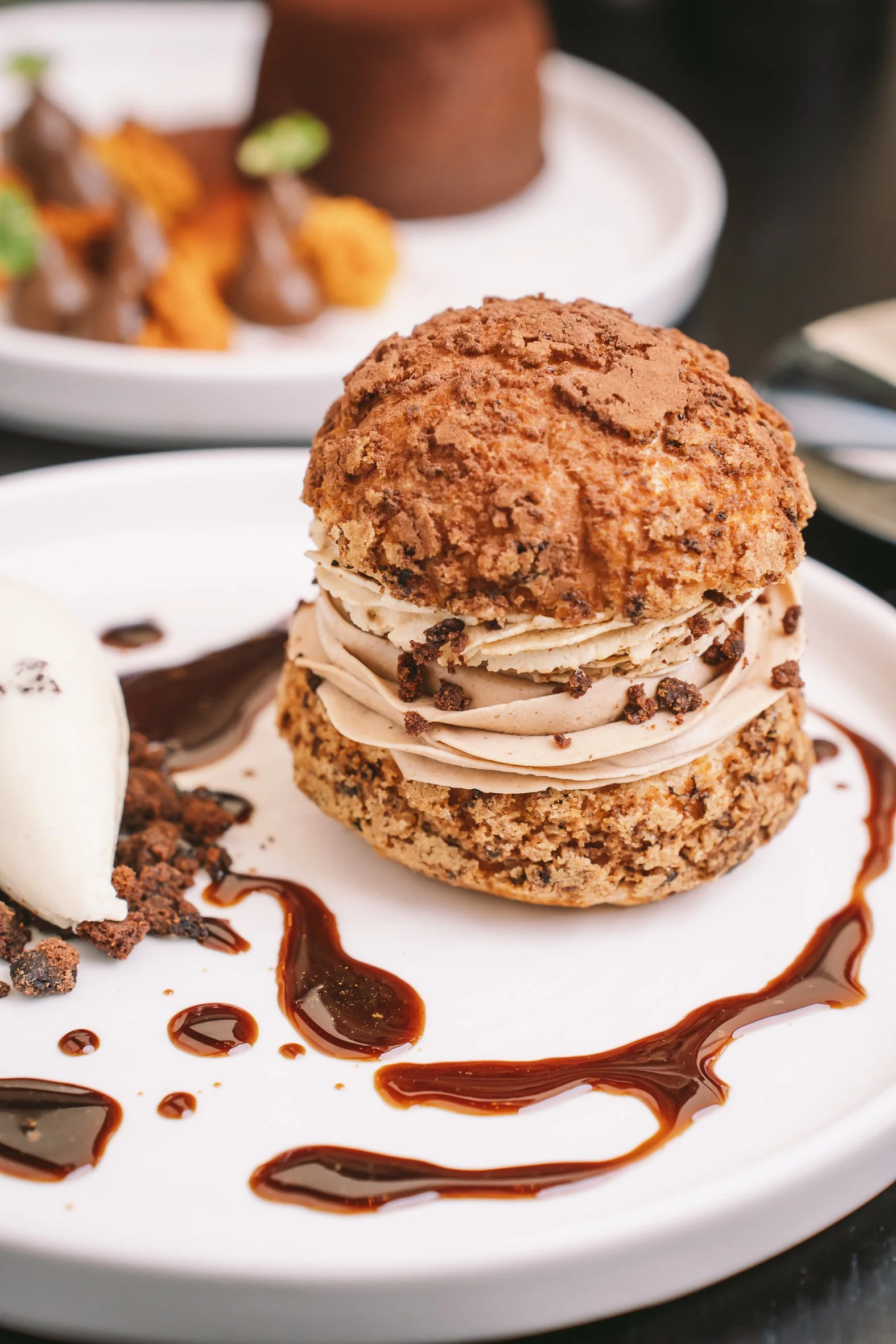 A dessert with a scoop of vanilla ice cream, a chocolate chip cookie topped with a cream swirl, and a cookie crumble on a white plate decorated with chocolate sauce.
