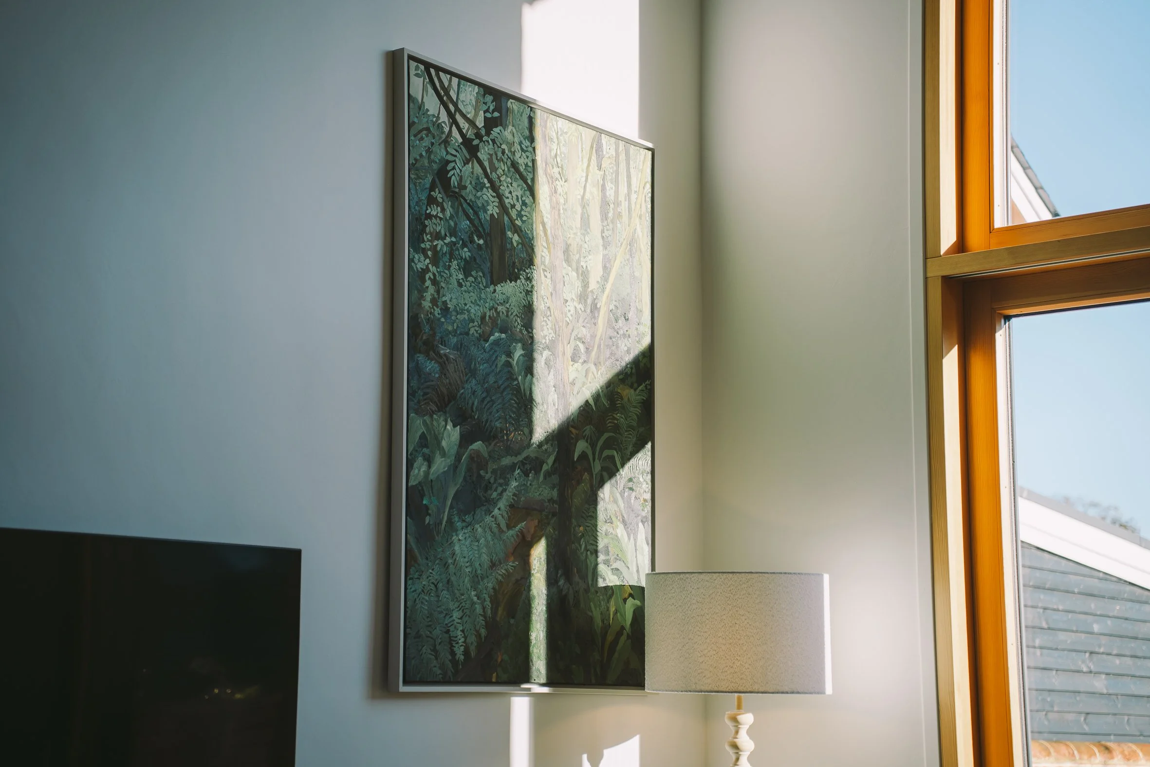 A framed painting of a lush green forest hangs on a white wall near a large window with orange trim, through which blue sky and rooftops are visible.