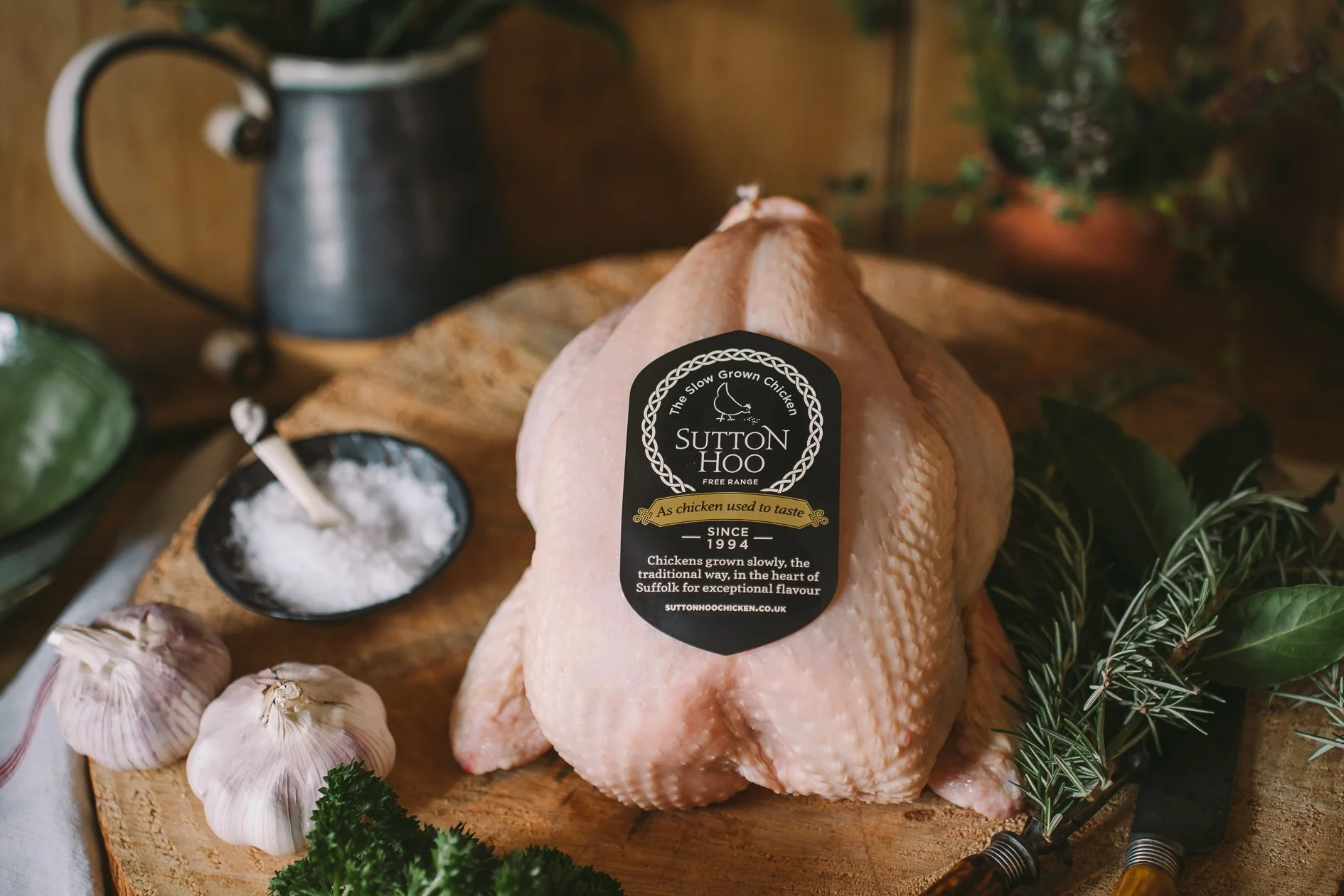 Raw whole chicken on a wooden cutting board with a label that reads 'Sutton Hoo, free range, since 1994, chickens grown slowly for exceptional flavor,' surrounded by garlic bulbs, fresh herbs, a small bowl of salt, a vintage-style metal watering can,