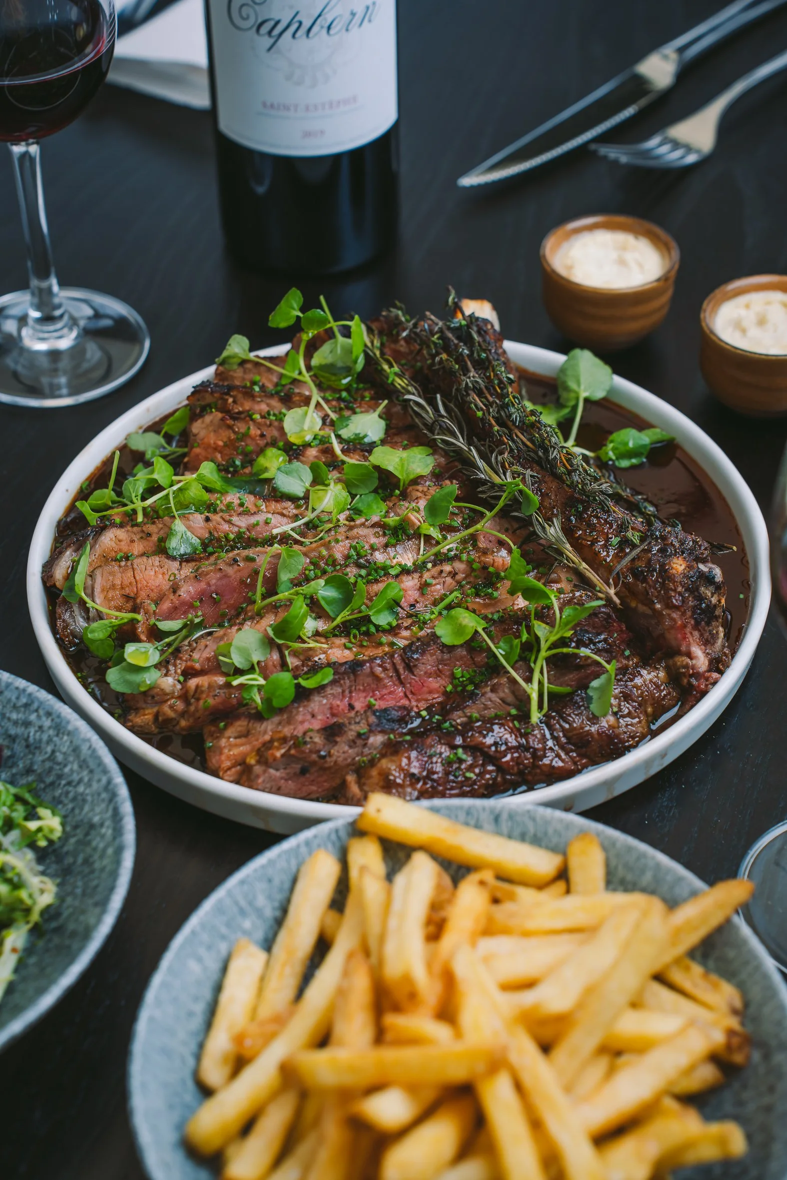 Close-up of a cooked meat dish garnished with fresh greens, with French fries on a plate in the foreground and a glass of red wine, a bottle of wine, and small bowls of dipping sauces on a dark table in the background.