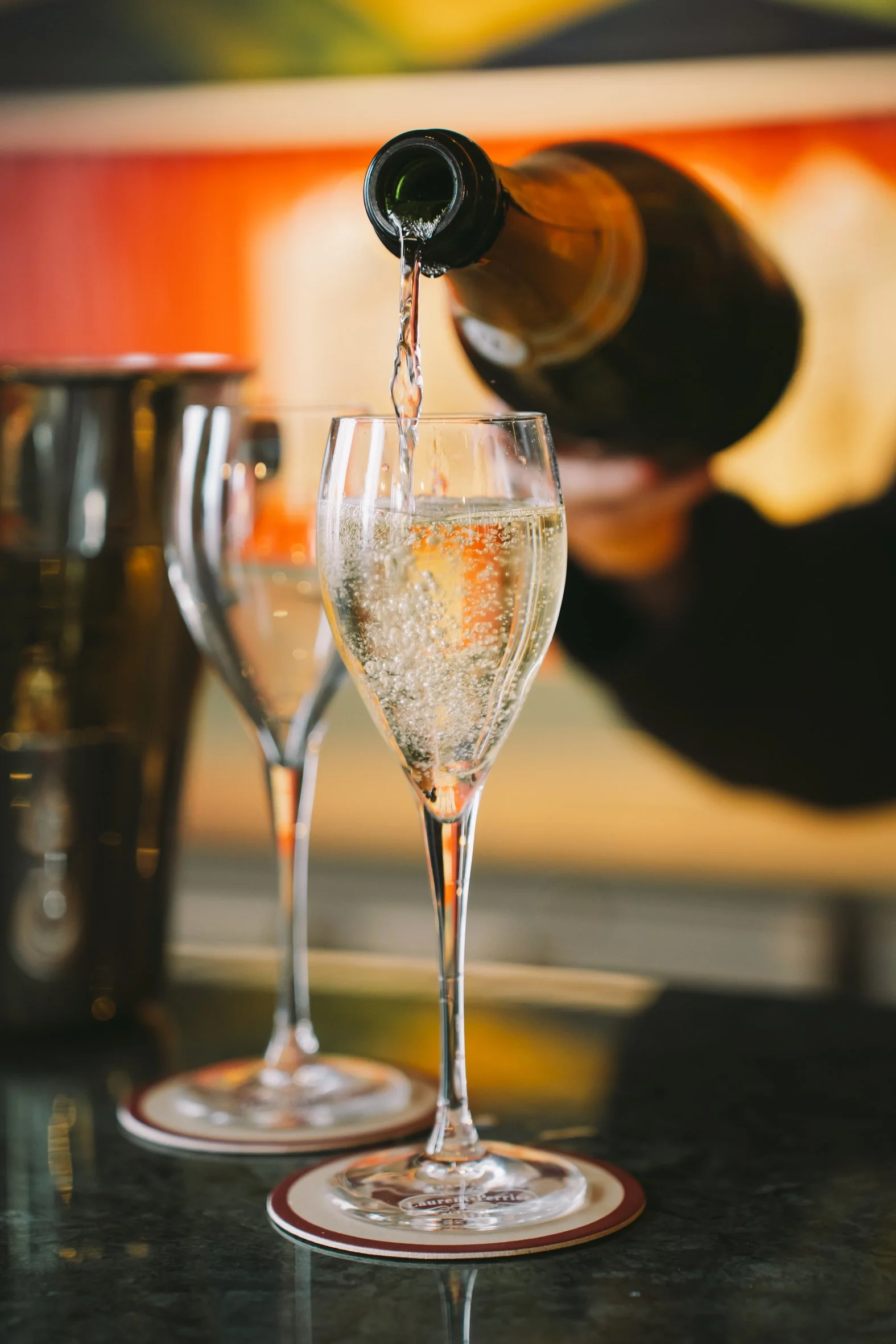 A bottle of sparkling wine is pouring into a champagne flute, with other glasses and a bottle in the background.