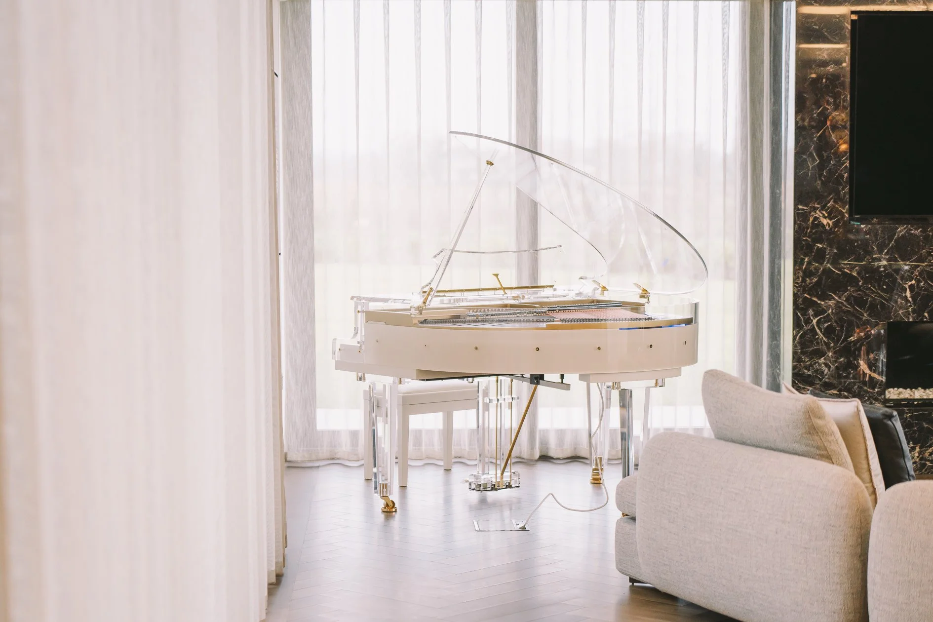 A white grand piano with a transparent lid and gold accents in a bright, modern living room with light-colored furniture, large windows with sheer curtains, a black marble fireplace, and hardwood flooring.