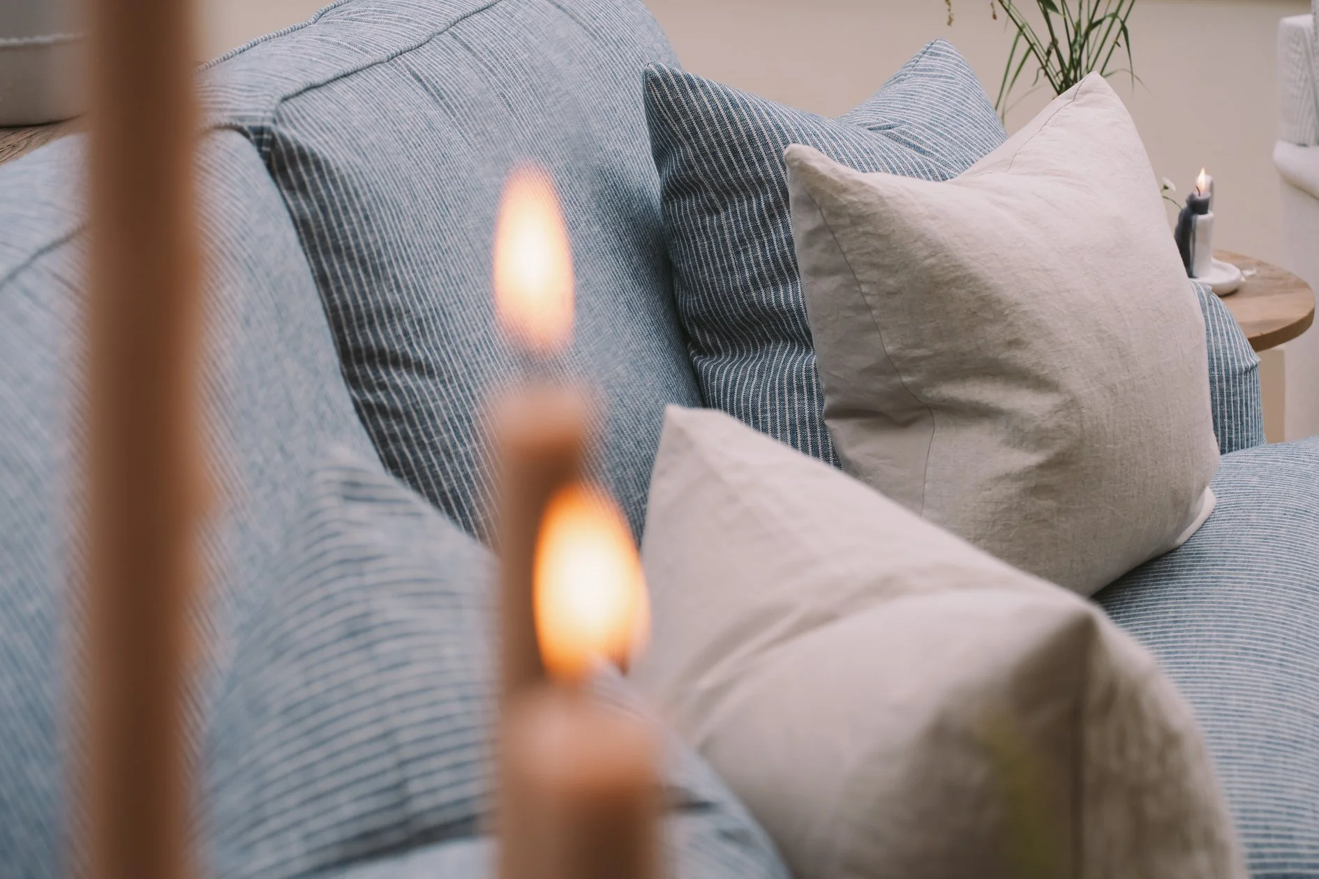 Close-up of a cozy living room corner with a blue striped sofa, beige cushions, a wooden side table with a candle, and plants, with soft lighting creating a warm ambiance.