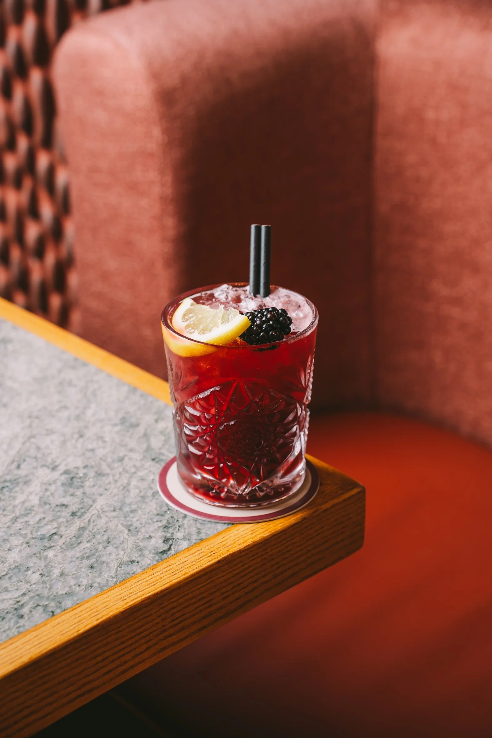 Close-up of a glass of red cocktail with lemon wedge, blackberries, and two black straws on a wooden table in a cozy setting.
