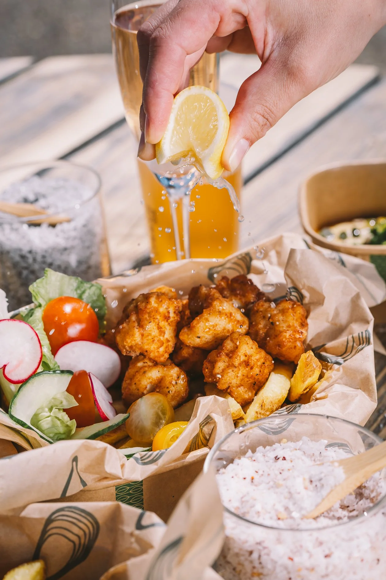 A person squeezing a lemon over a plate of fried chicken with vegetables, salad, and sides at a outdoor meal.