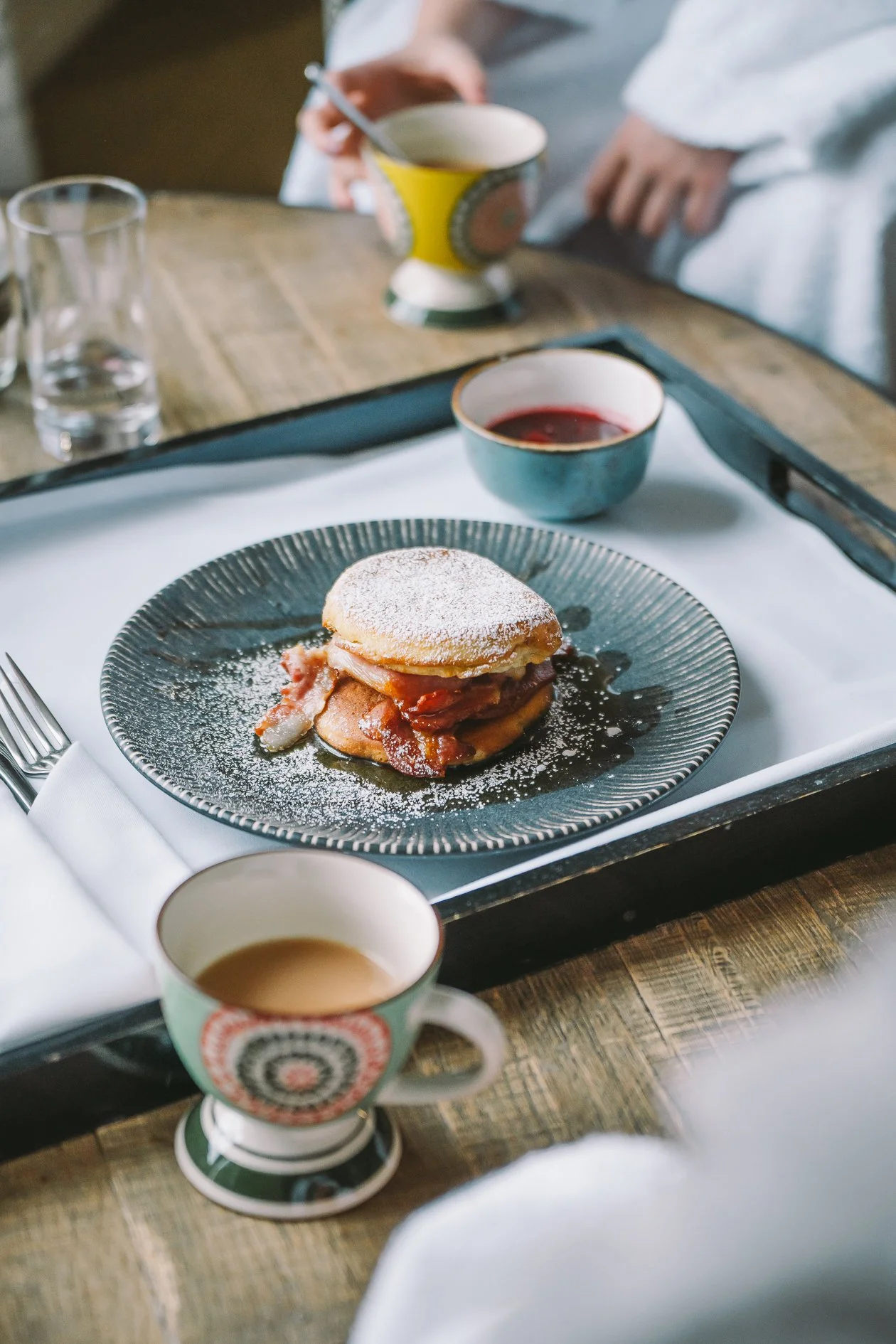 A breakfast tray with a plate of eggs benedict, a small bowl of sauce, a cup of coffee, a glass of water, and a person in the background holding a mug.