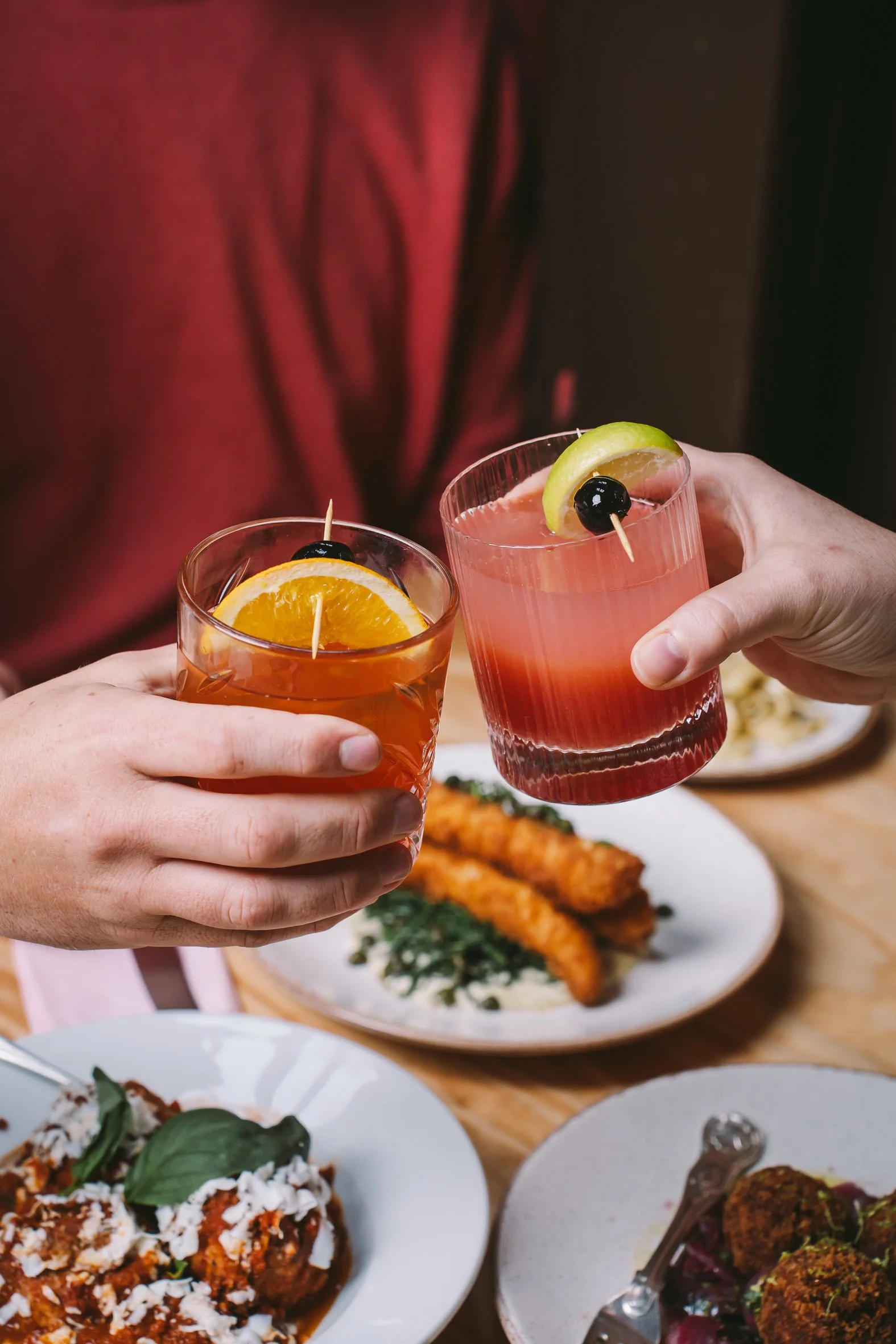 Two people holding drinks with garnishes, sitting at a table with plates of food, including fried items and pasta.