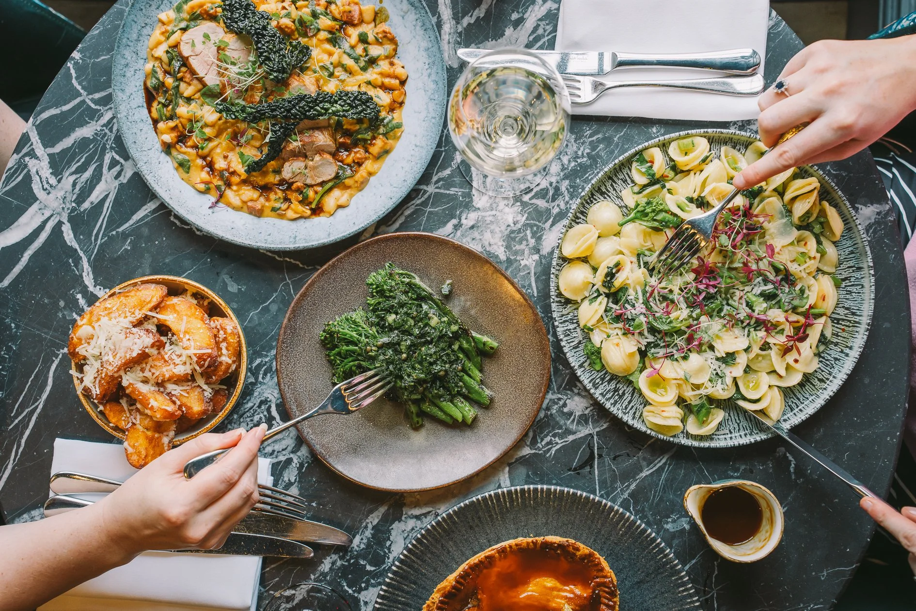 A top-down view of a dinner table with various dishes, including pasta with greens and microgreens, a plate of mashed potatoes with gravy, grilled broccoli, and another dish that appears to be a baked pasta or casserole. There is a glass of white win