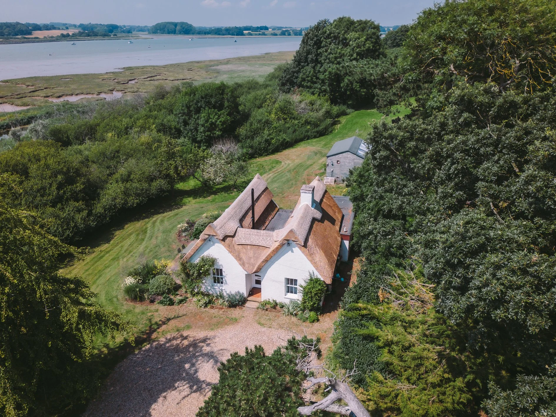 Aerial view of a white house with a thatched roof surrounded by green trees and a gravel driveway, with a river in the background.