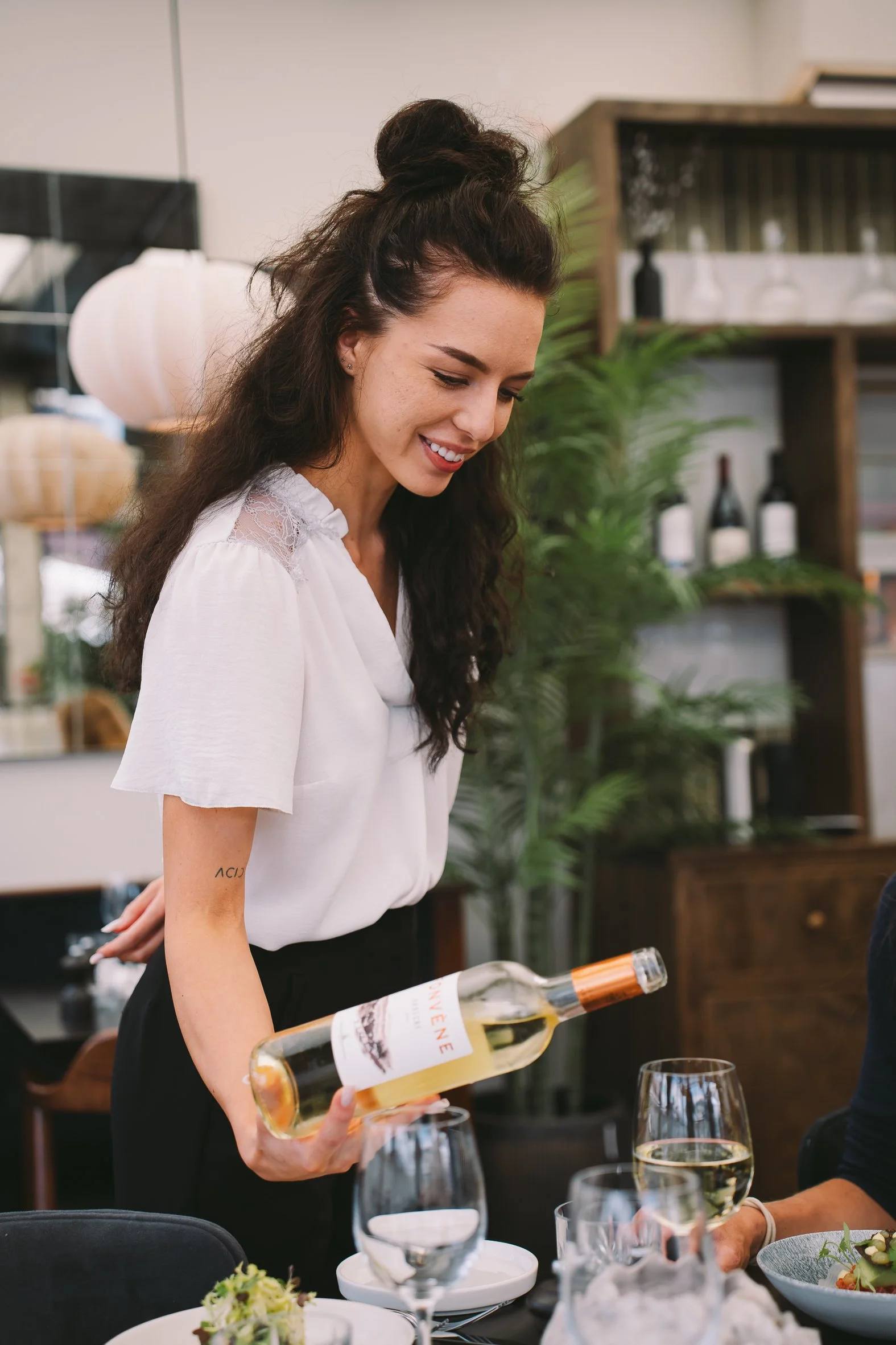 A woman in a white blouse and black skirt is pouring white wine into a glass at a dinner table set with multiple wine glasses, plates, and bowls, with other people and decorative plants in the background.