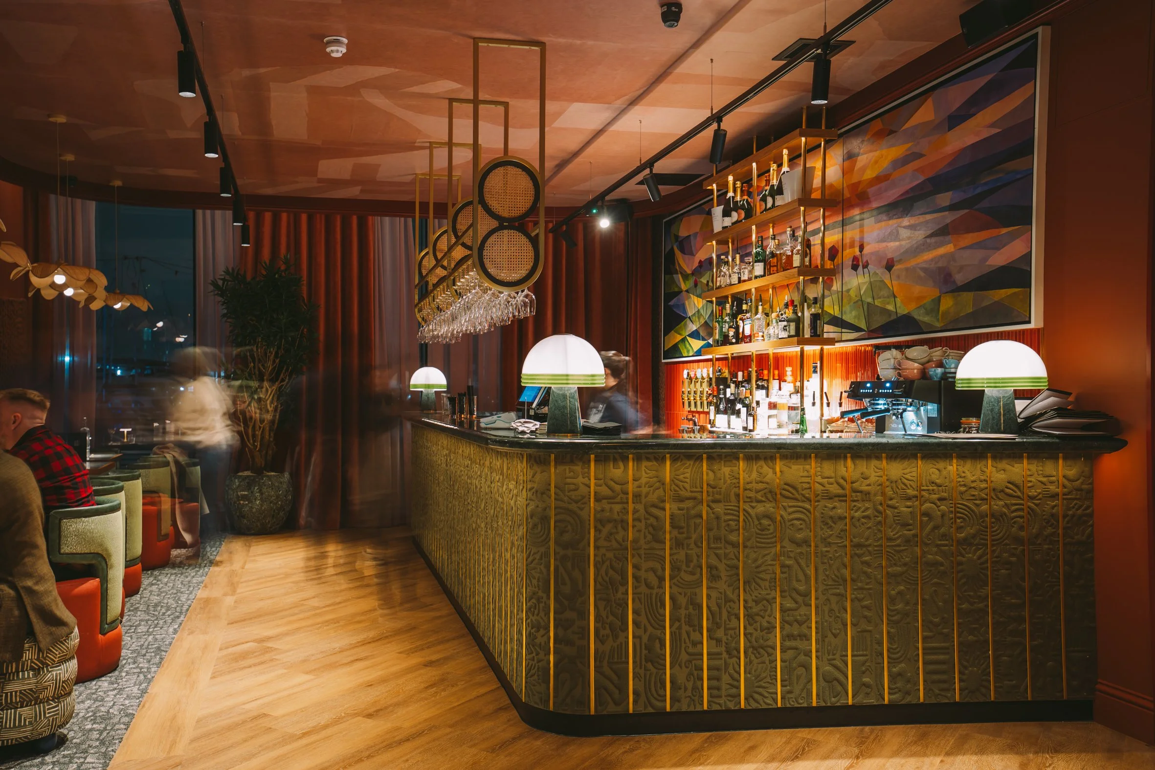 Interior of a bar with a curved wooden counter, modern pendant lights, shelves with bottles of alcohol, and colorful abstract wall art.