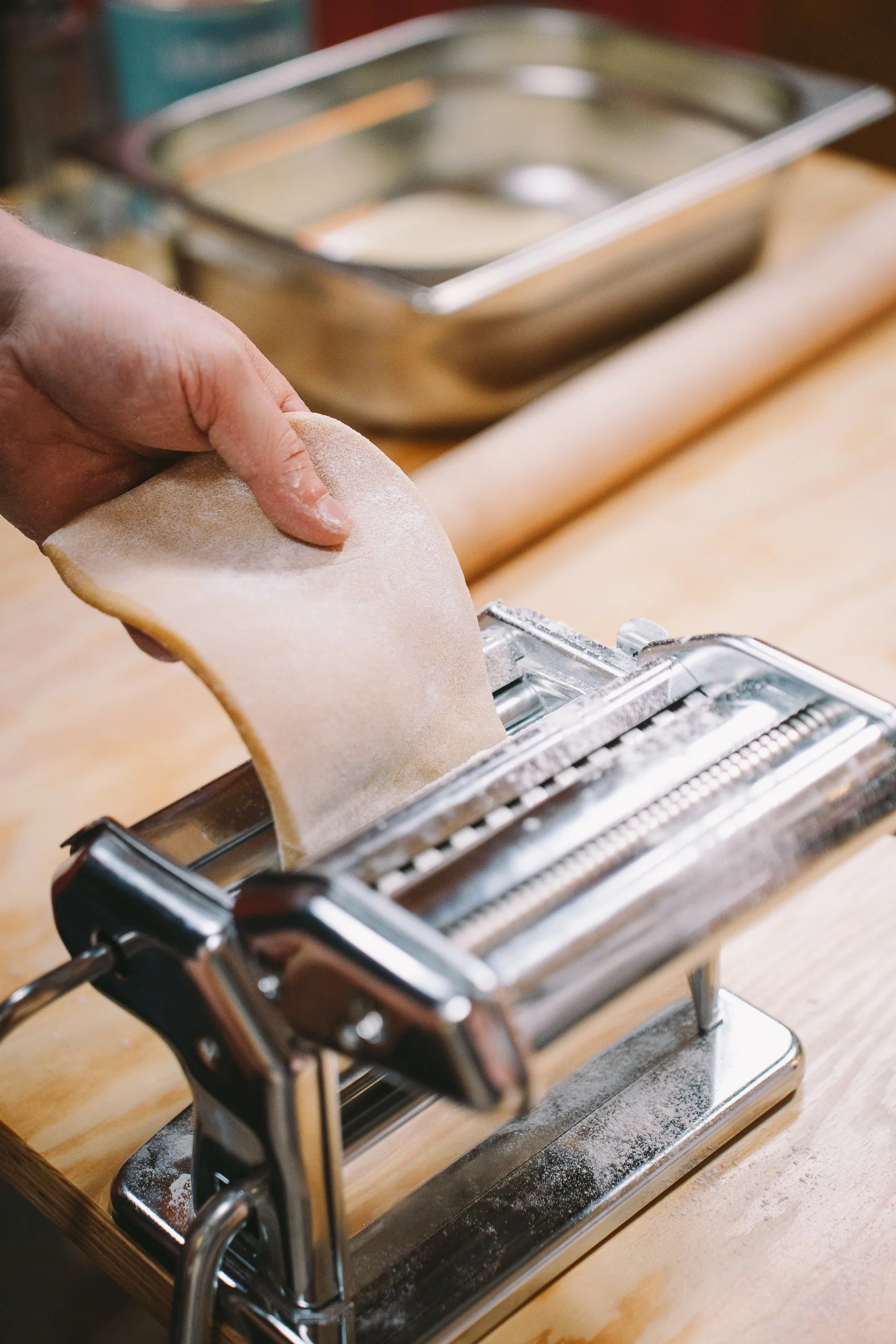 A person's hand feeding a piece of uncooked pasta into a silver pasta cutter on a wooden surface.