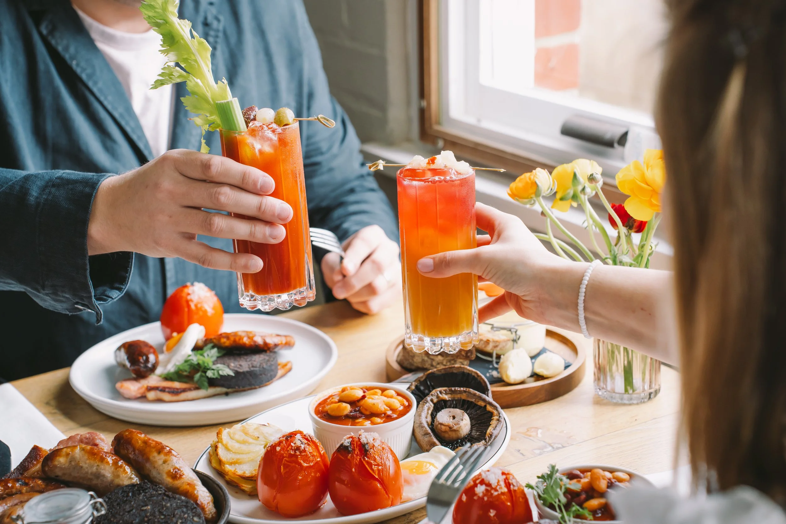 People enjoying a meal with bright orange cocktails garnished with celery and olives, surrounded by plates of food including tomatoes, mushrooms, and various appetizers on a wooden table near a window with a vase of yellow flowers.