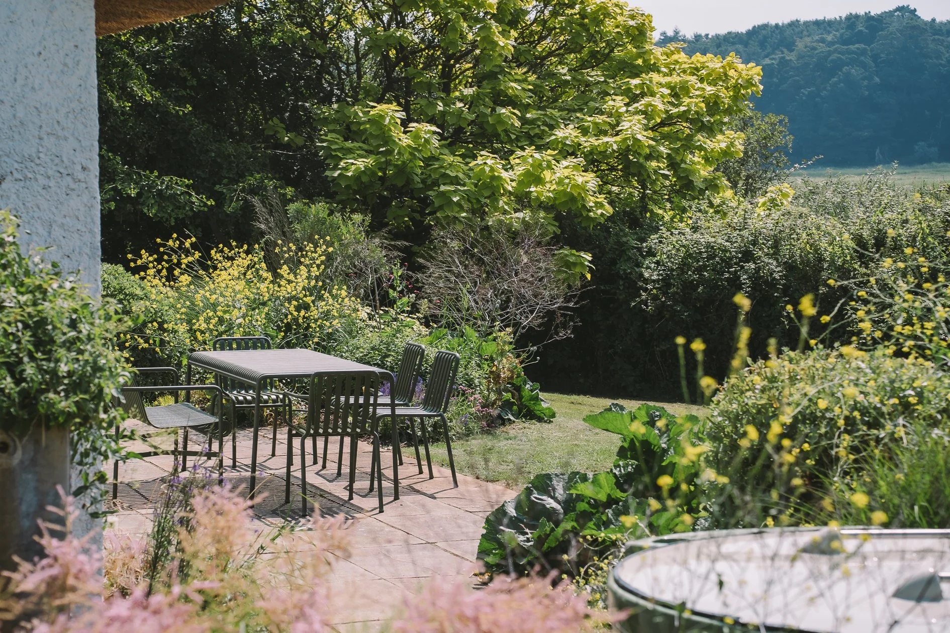 Outdoor garden with patio table and chairs surrounded by lush green plants and trees, with a grassy yard and distant hills in the background.