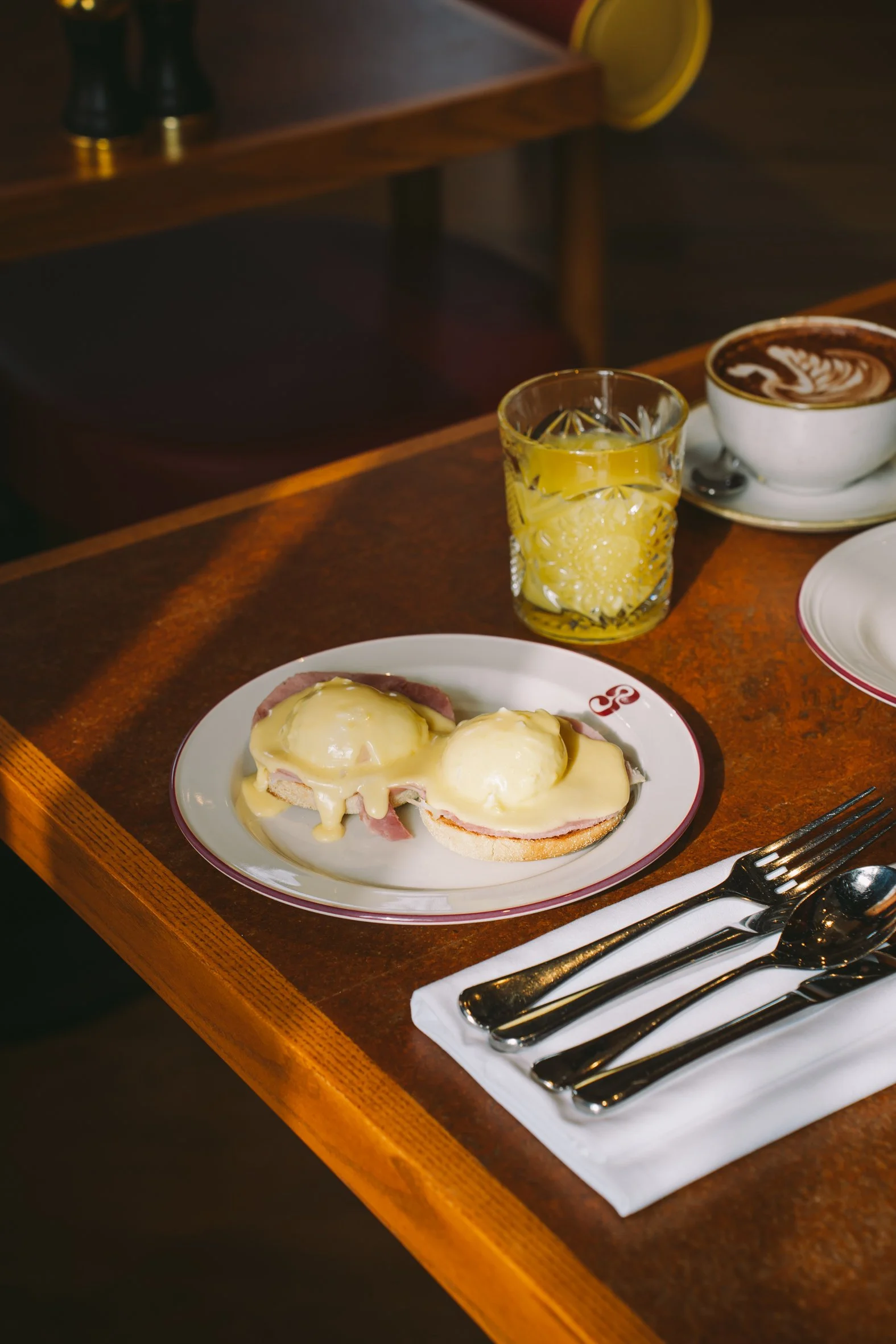 A plate with eggs benedict topped with hollandaise sauce, a glass of orange juice, and a cup of coffee on a wooden table.