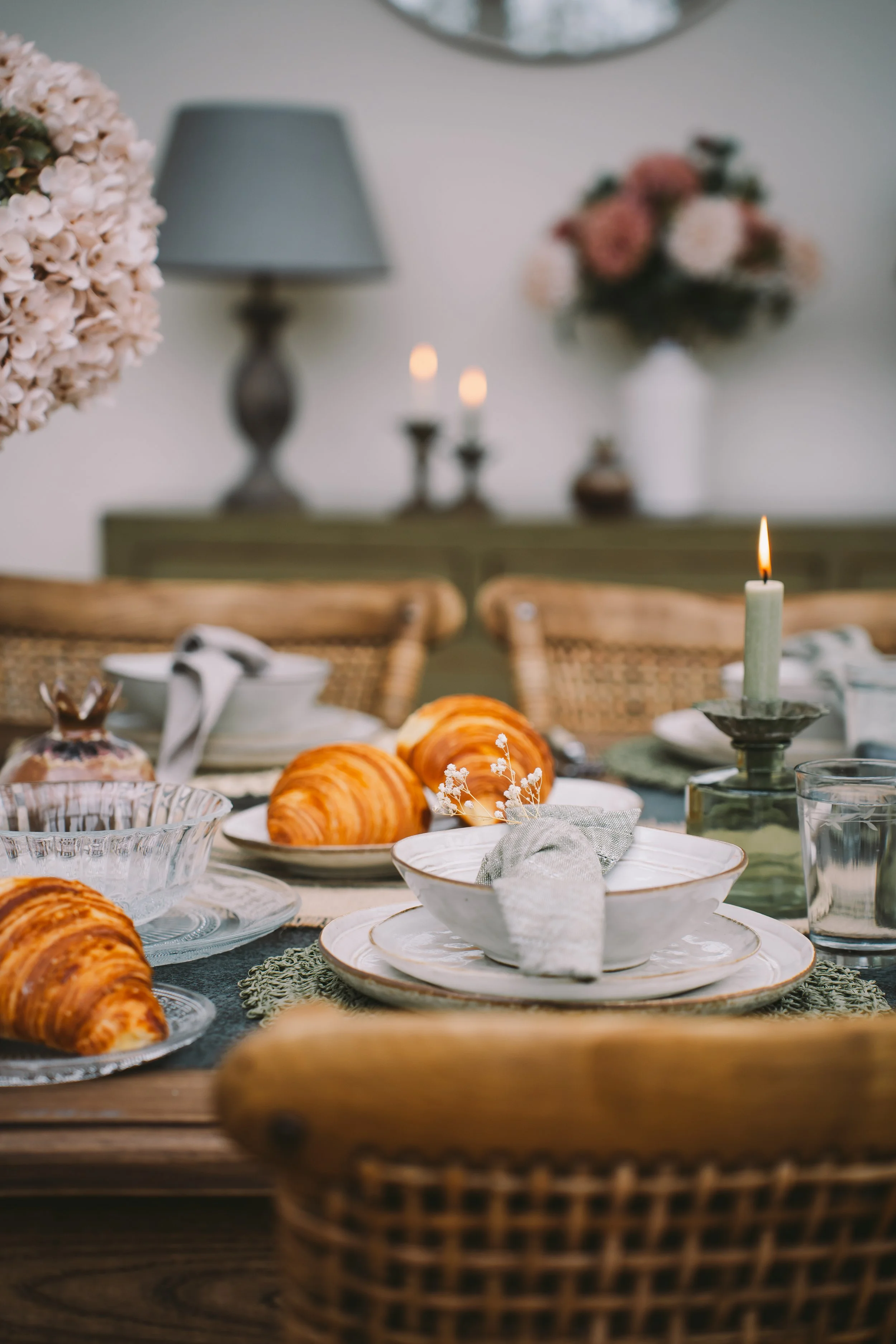 A cozy dining table set with white plates, a cloth napkin, glassware, and fresh croissants. The background features a blurred interior with floral arrangements, a lamp, and candles, creating an inviting atmosphere.