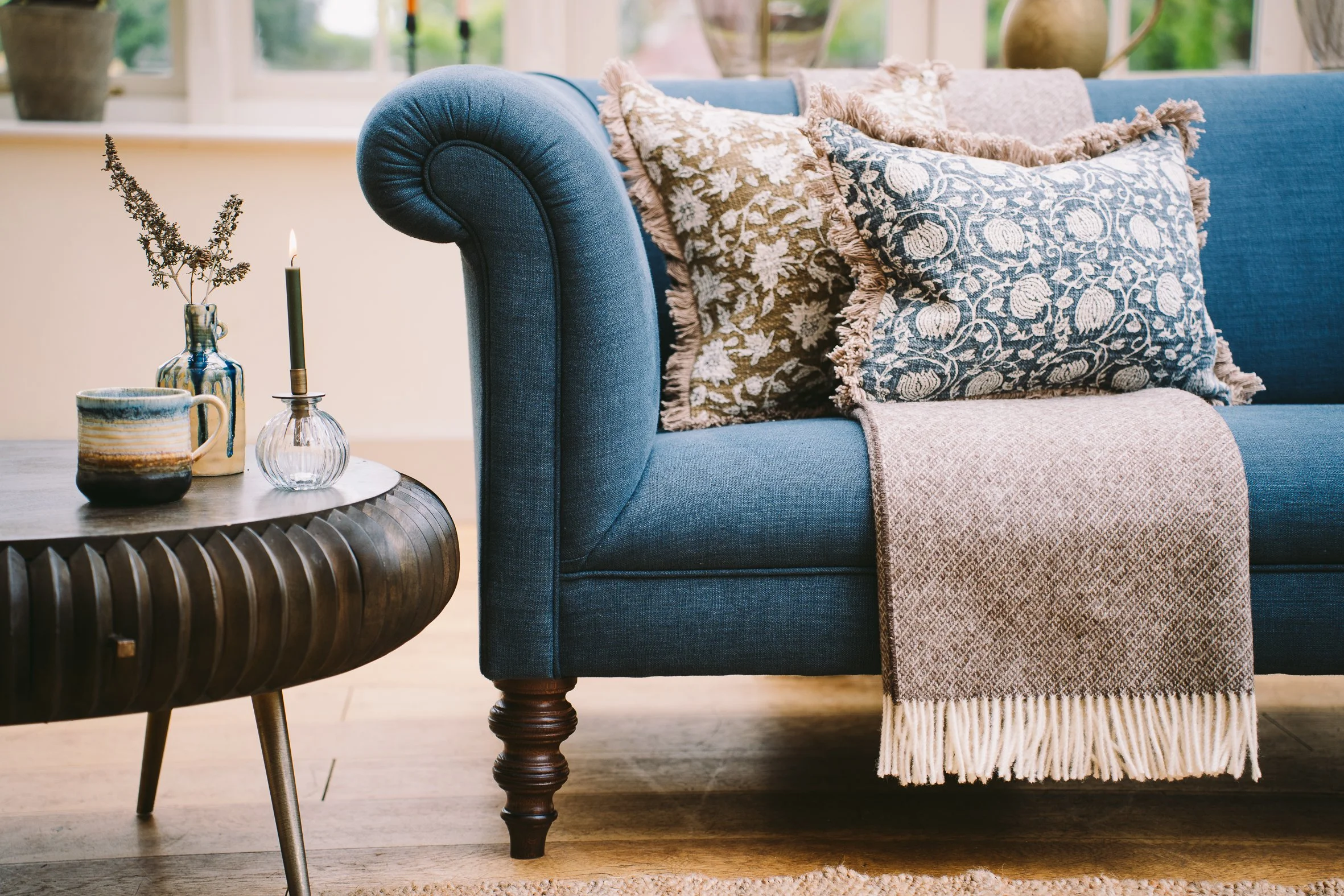 A cozy living room scene featuring a blue upholstered sofa with decorative pillows and a beige and blue throw blanket. A wooden side table holds a candle and decorative vases with dried plants, set before a large window with natural light.