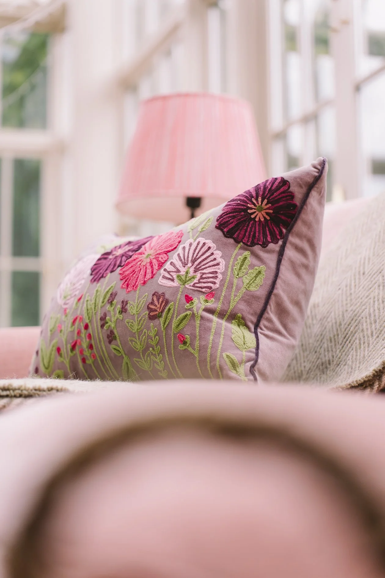 A pink embroidered pillow with floral patterns, including large dark purple and pink flowers and green leaves, placed on a sofa in a sunlit room with large windows.