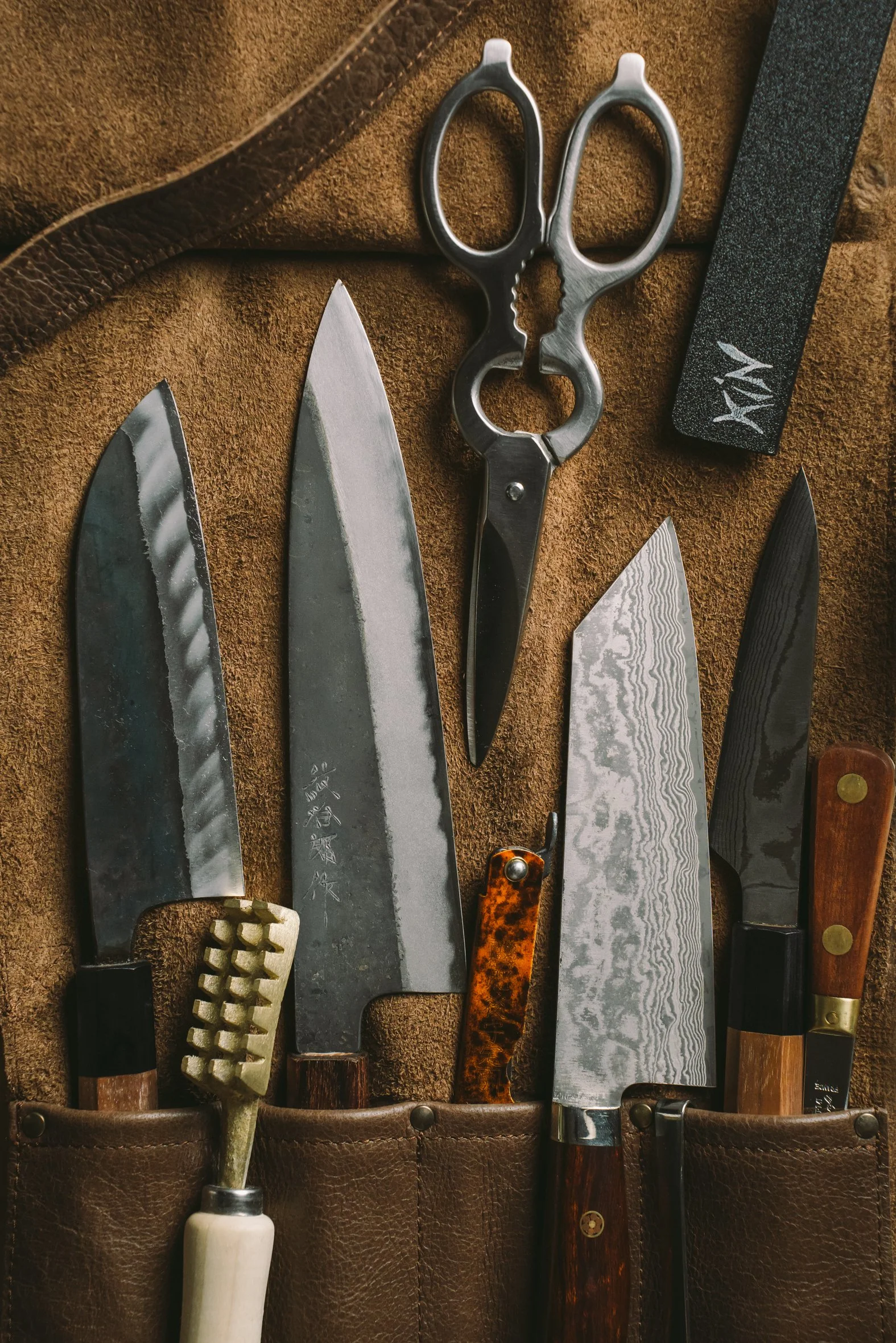 Collection of various knives and scissors stored in a leather knife roll, including a white-handled kitchen tool, a small carving knife with a tortoise shell handle, and several chef's knives with different blade patterns.