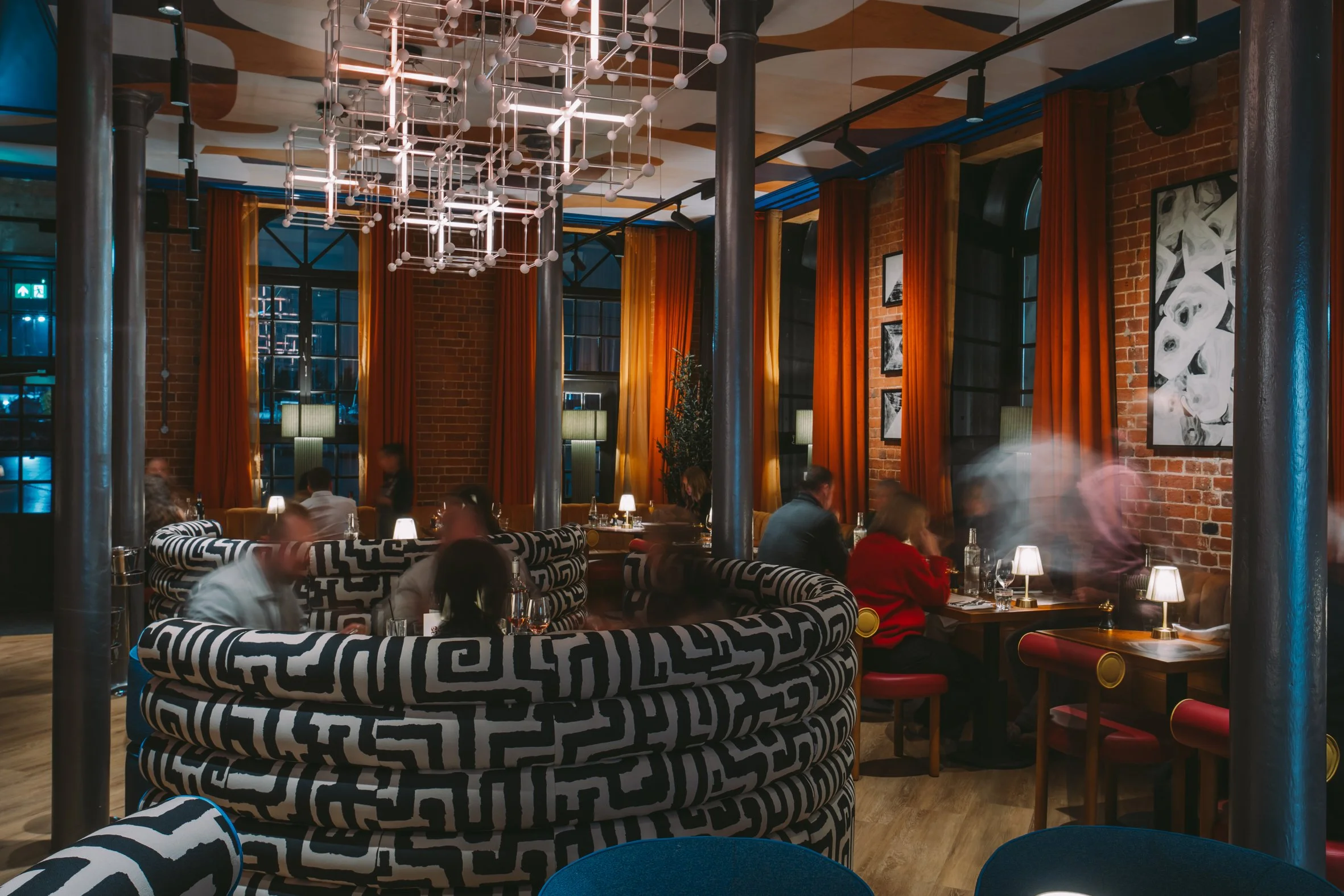 Interior of a modern restaurant with exposed brick walls, long orange curtains, black and white maze-patterned cushioned seating, hanging geometric chandelier, and several patrons dining.