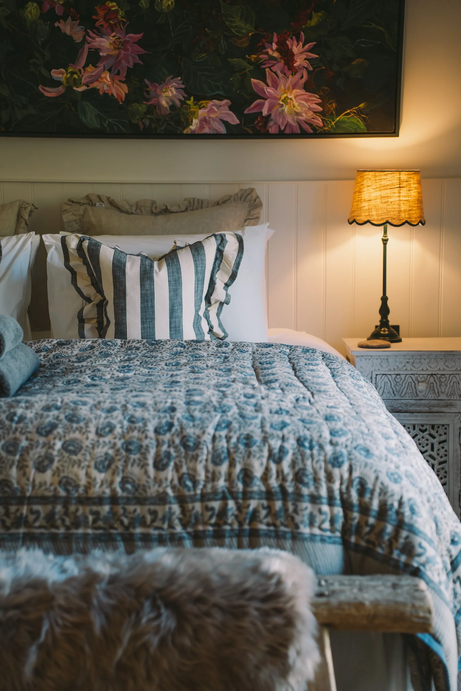Close-up of a fluffy light-colored dog in front of a bed with pillows and a quilt, with a table lamp and a framed floral painting on the wall behind.