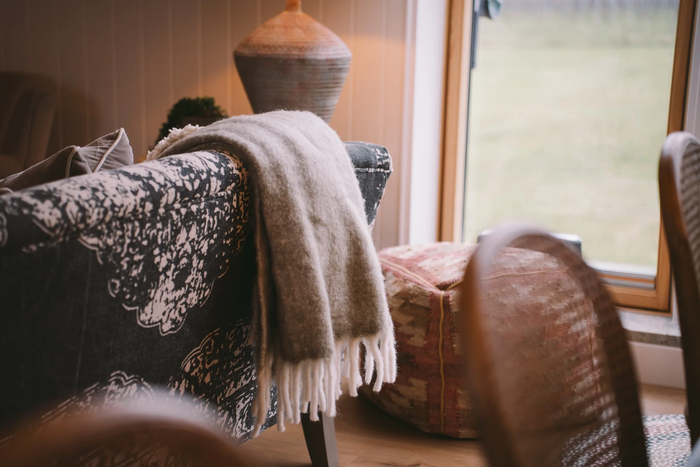 Close-up of a cozy room corner with a patterned armchair, a throw blanket, a cushion, a large decorative vase, and a window showing an outdoor view.