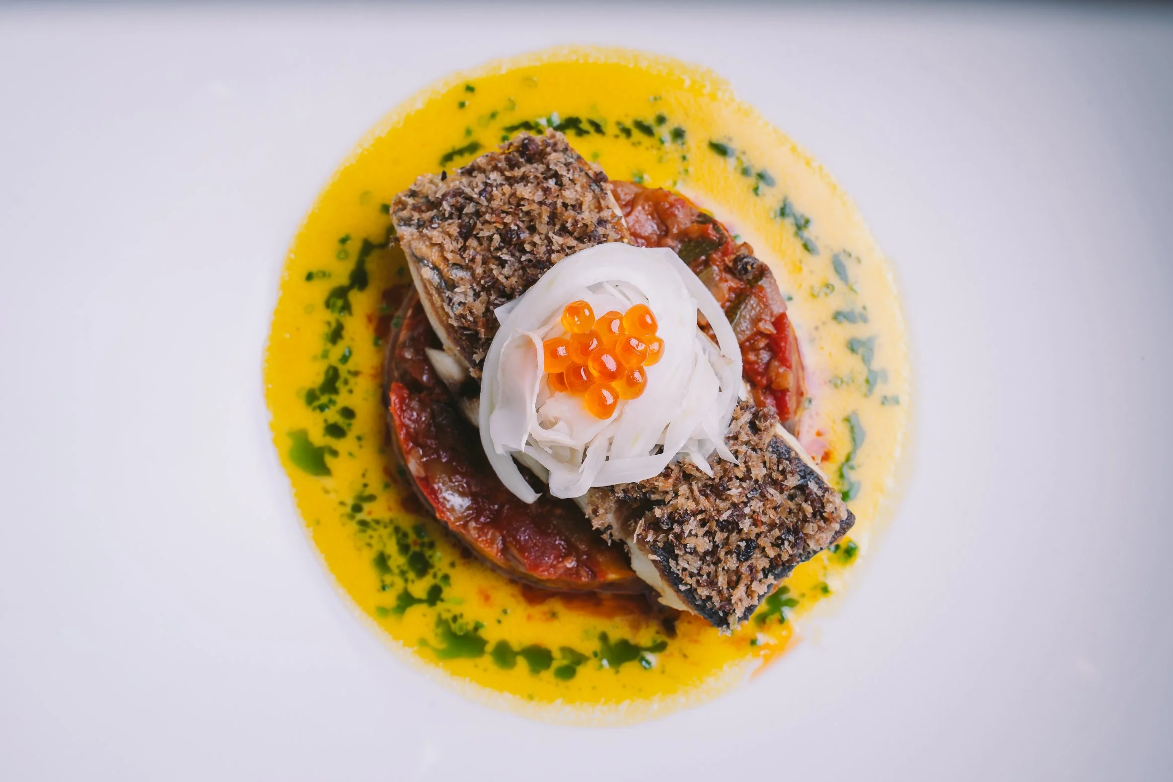 A plated gourmet dish featuring a slice of meatloaf with a crust, topped with a dollop of sour cream, orange fish roe, and a slice of onion, all on a bed of tomato sauce, with a yellow sauce and green herb garnish surrounding the plate.