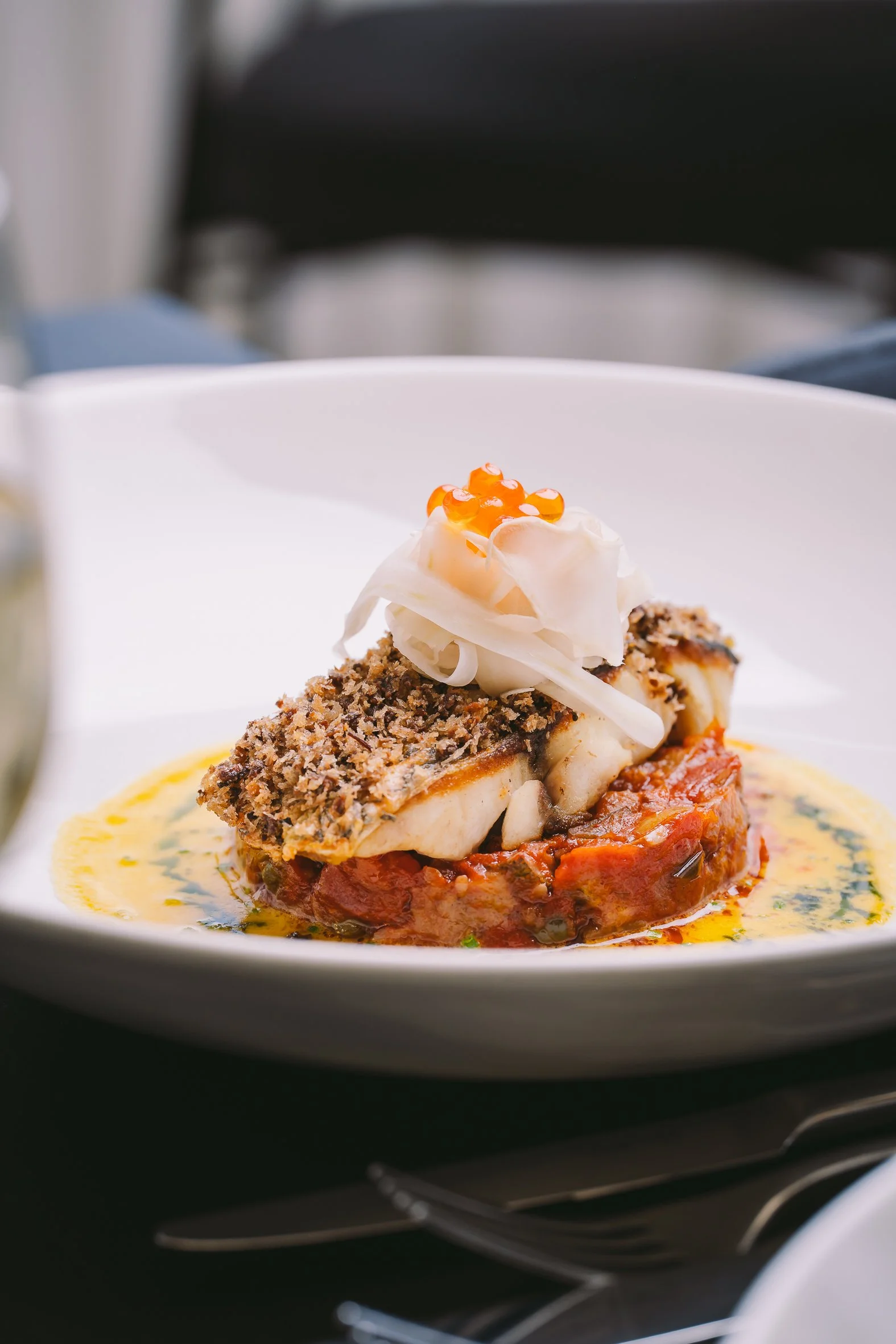 A plated fish dish with a crumb coating, topped with coconut flakes and orange caviar, served on a bed of tomato or red pepper sauce, garnished with a creamy sauce and herbs.